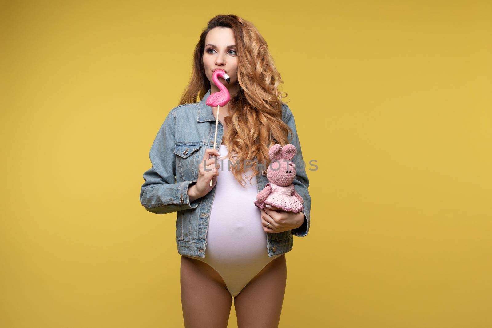 Funny young casual pregnant girl posing with hare toy and sweet candies isolated at yellow studio background. Beautiful fashion pregnancy woman enjoying motherhood medium long shot