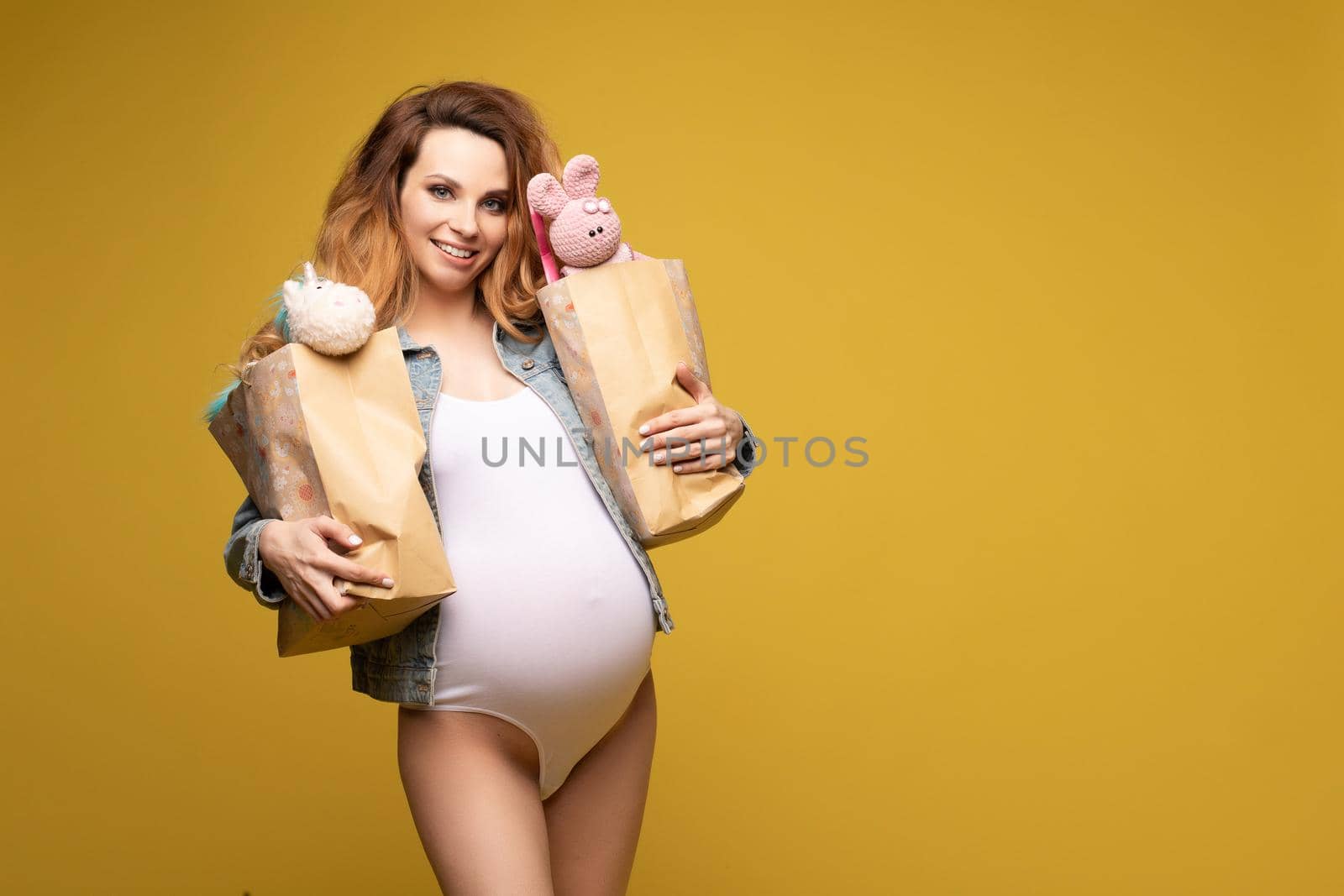 Waist up of smiling pretty pregnant female wearing in white bodysuit and denim jacket on yellow background
