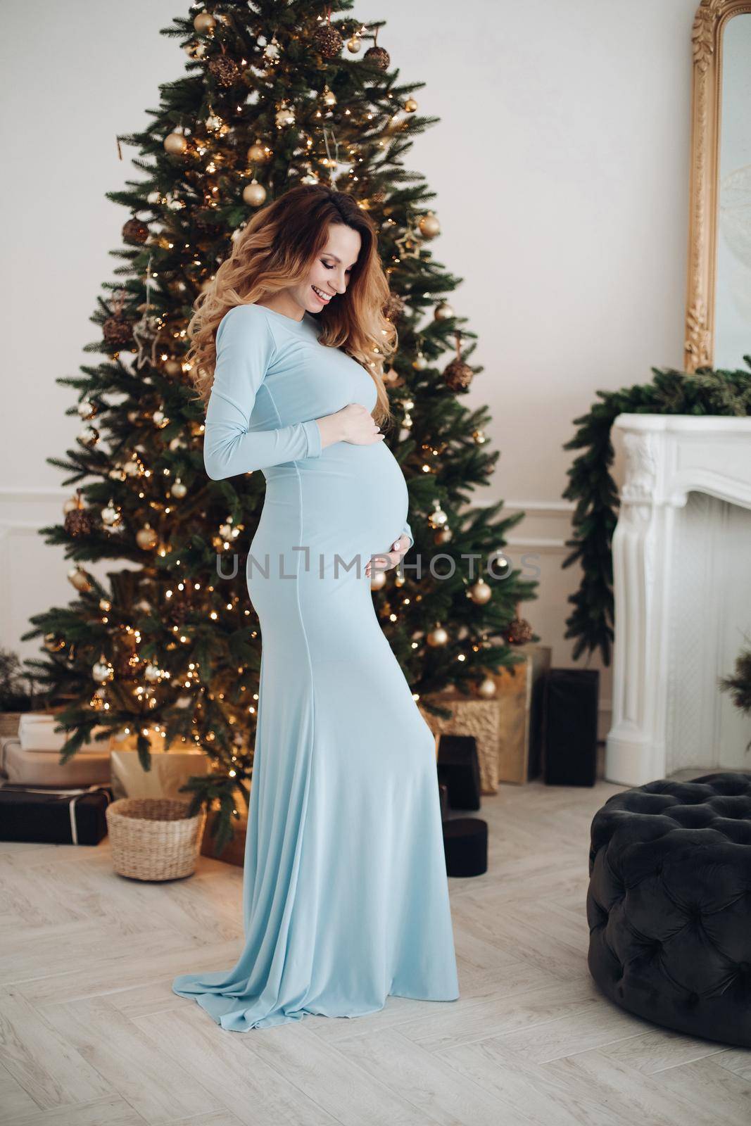 Full length of pregnant female in blue dress smiling joyfully in studio on the background of New Year lights. Christmas and New Year concept