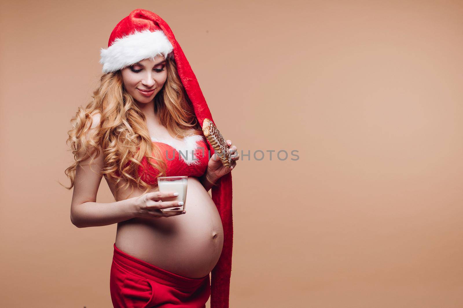 Smiling beautiful pregnant lady in Santa Claus hat soaking gingerbread in milk on beige background. Christmas and New Year concept