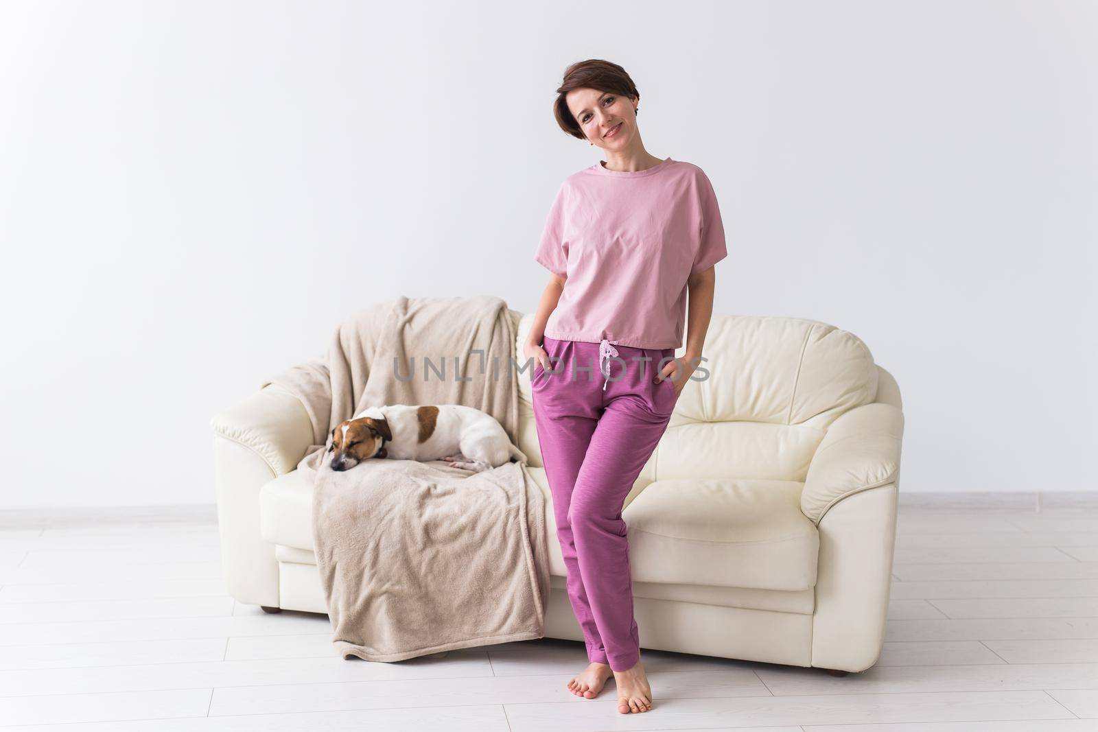 Young attractive woman dressed in beautiful colorful pajama posing with dog as a model in her living room. Comfortable sleepwear, home relaxation and female fashion concept. by Satura86