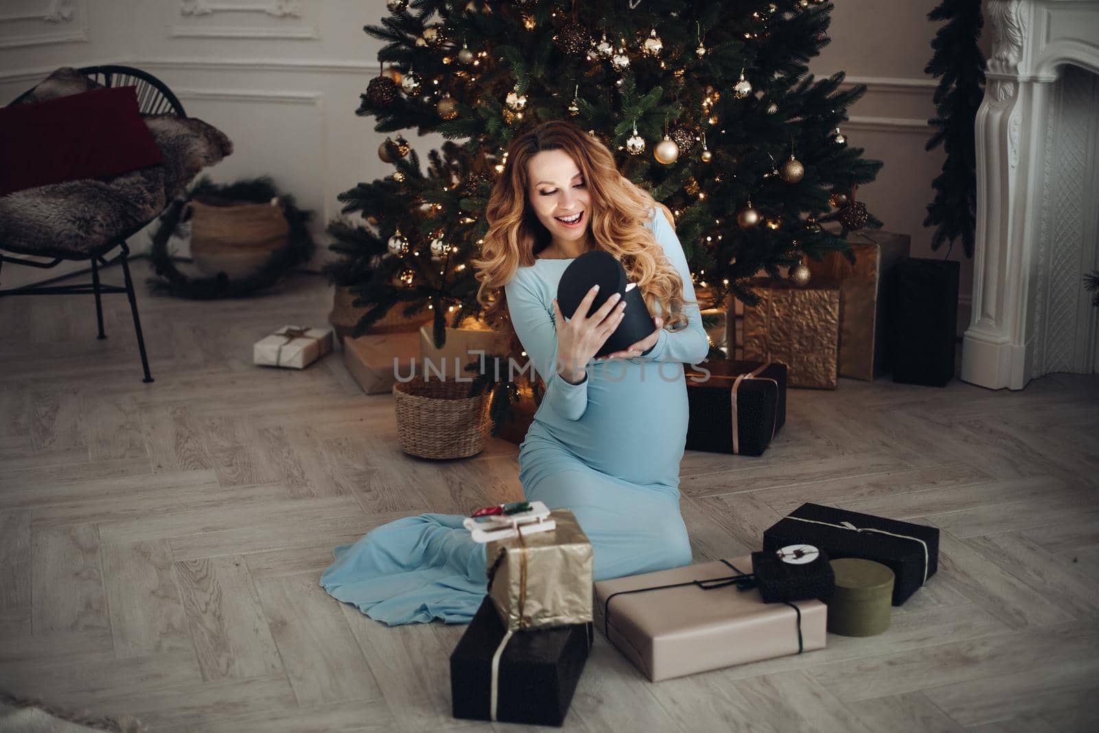 Waist up of smiling pretty pregnant lady opening present while sitting near Christmas tree. Christmas and New Year concept