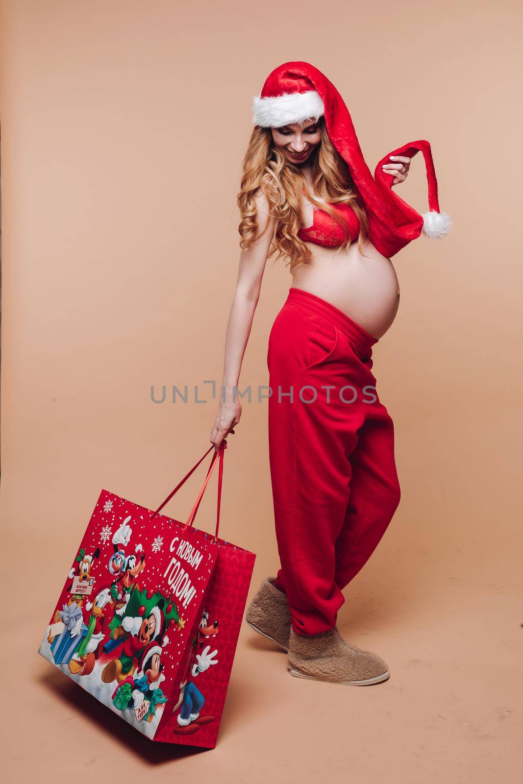 11 27 2019 Belarus Minsk: Happy funny Christmas pregnant girl in Santa Claus costume holding holiday gift having positive emotion. Smiling pregnancy female celebrating New Year season isolated at nude studio background