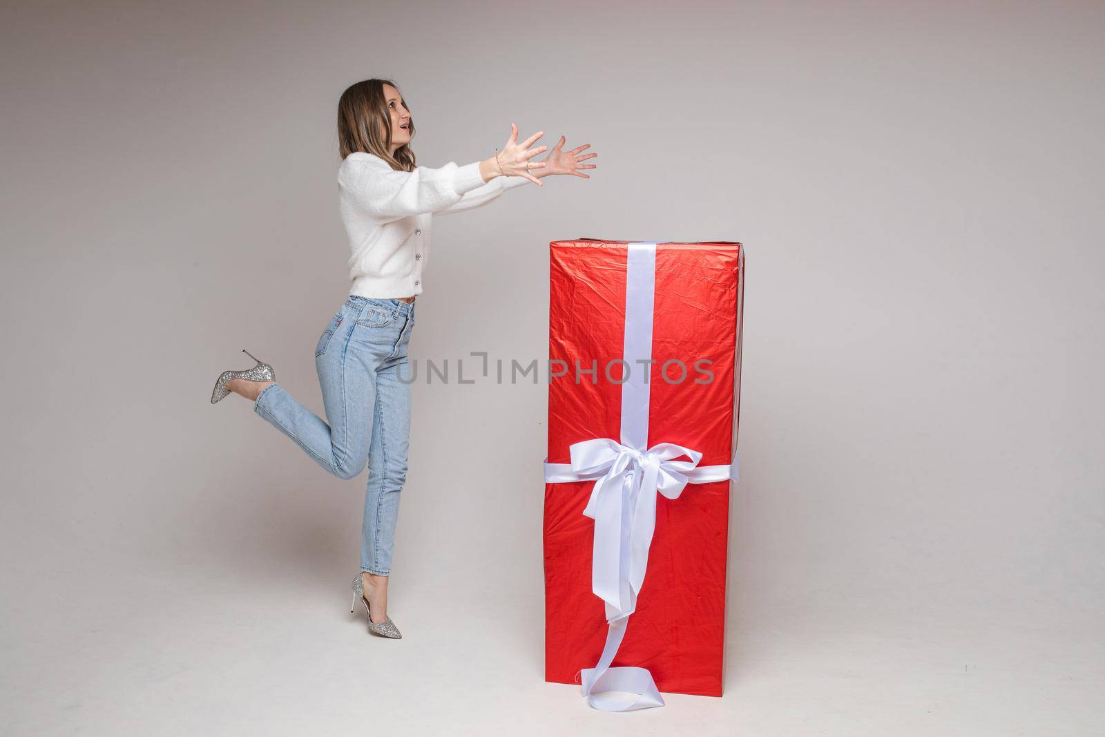 caucasian woman with attractive appearance rejoices of a big present for st. valentine's day and jumps near it, picture isolated on white background