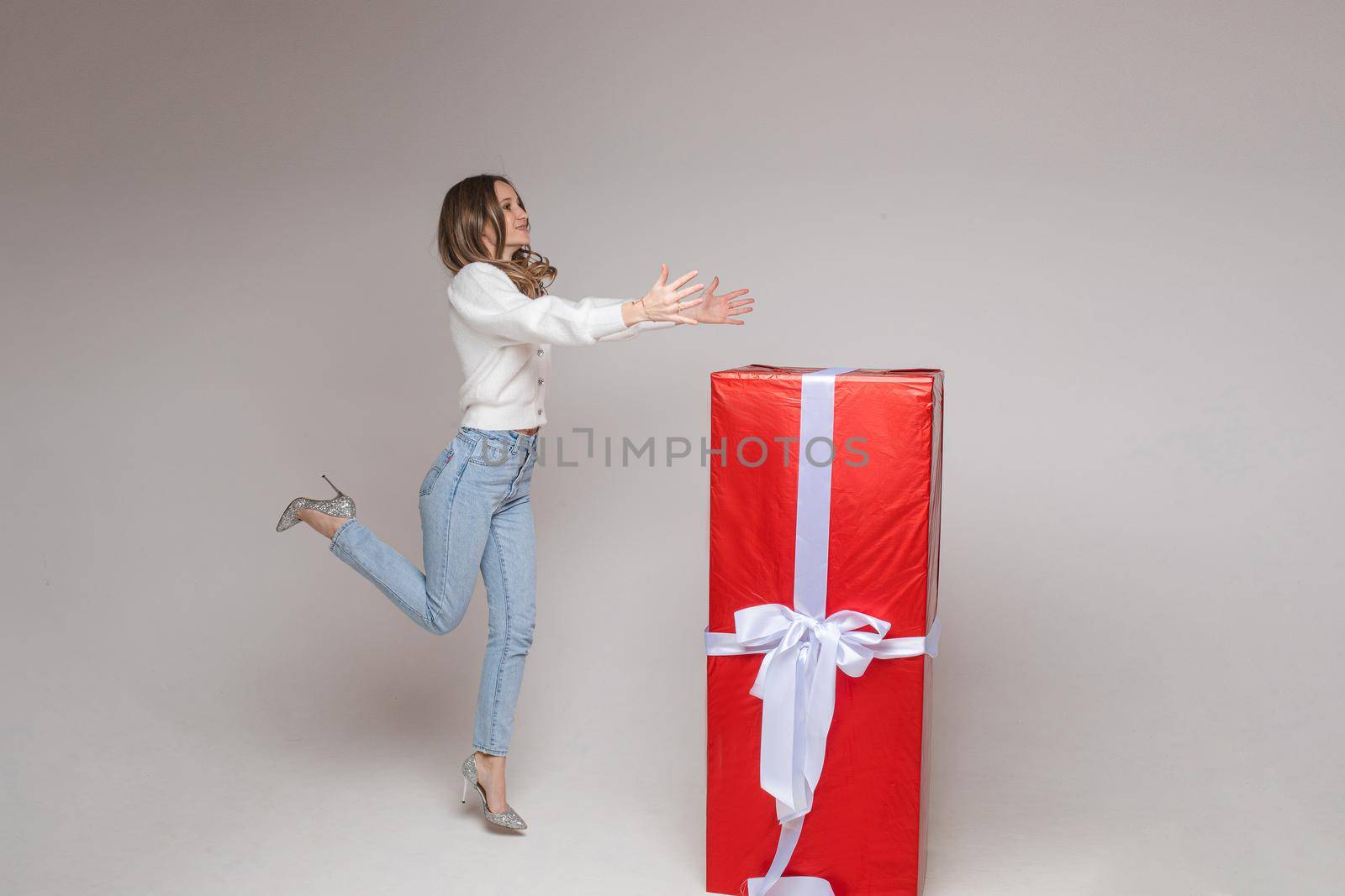 Full length stock photo of cheerful jumping girl in heels with outstretched arms with giant gift in red paper with white bow on white background.