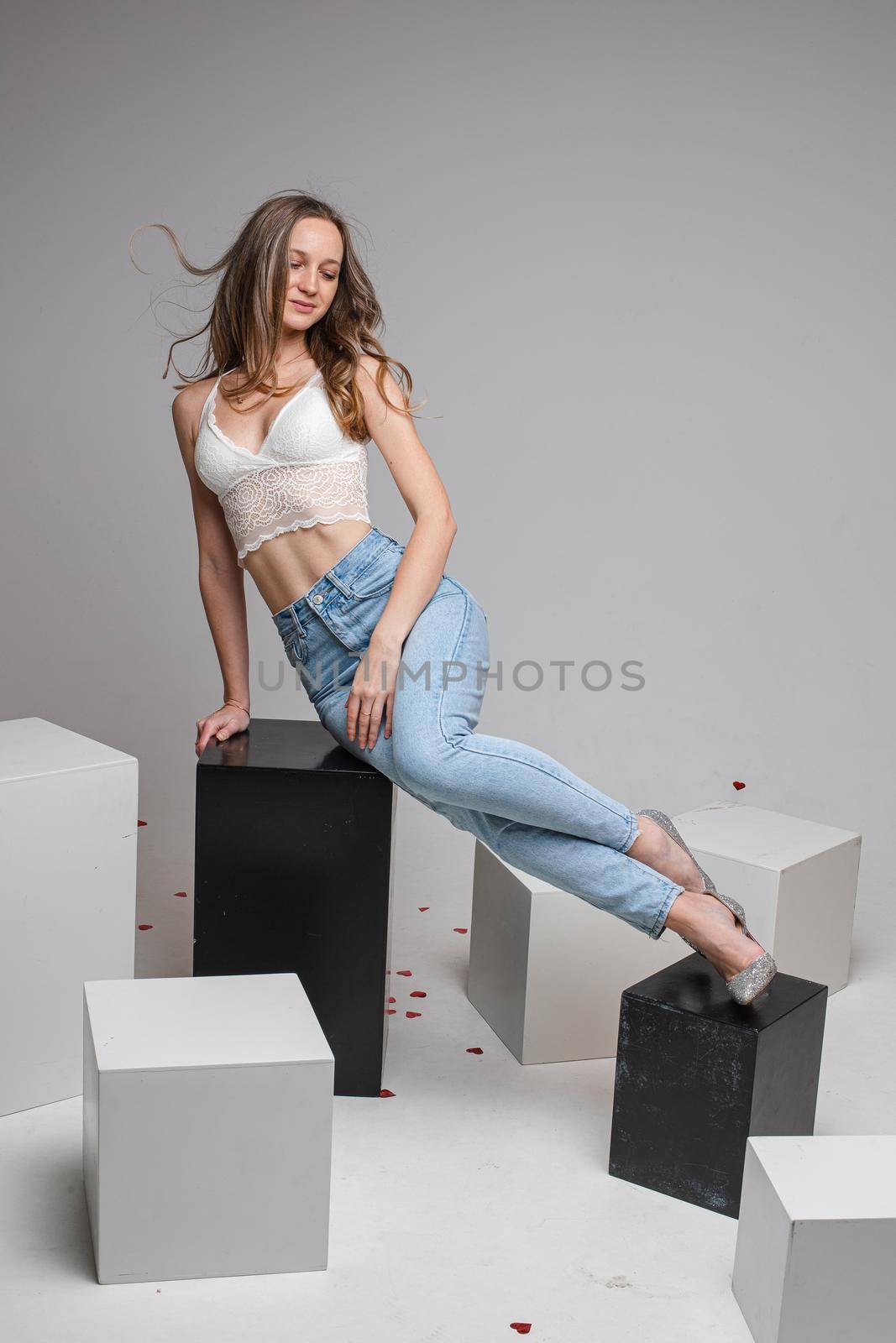 Pretty slim long haired caucasian girl in white top lingerie, blue jeans posing for studio portrait on gray background by StudioLucky