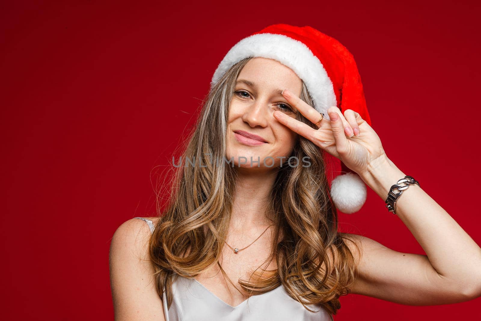 Portrait of jovial carefree girl with wavy fair hair wearing red Santa hat with white rim showing peace sign with fingers at eye and sticking her tongue out. Isolate on red.
