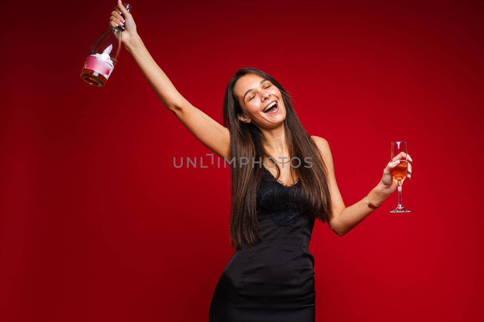 cheerful caucasian woman with attractive appearance with a bottle of champagne and glass, picture isolated on red background