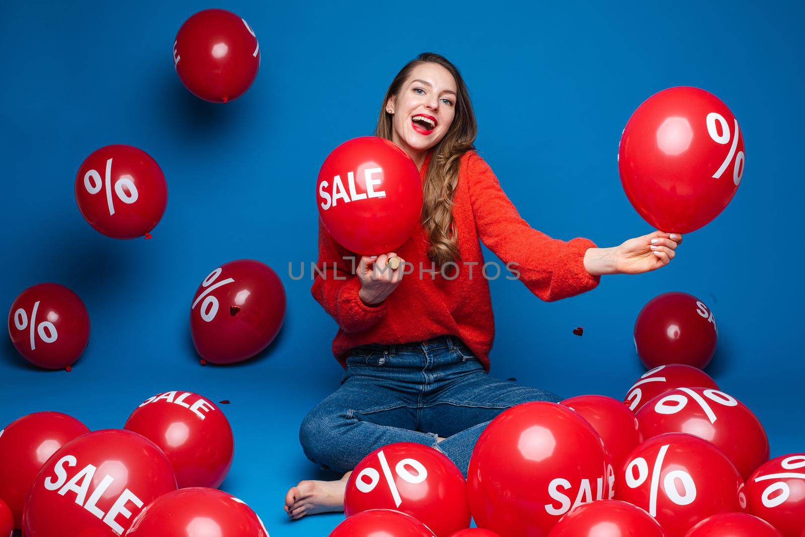 Joyful young girl sitting on the floor on blue studio background with red balloons, sale and discount concept by StudioLucky