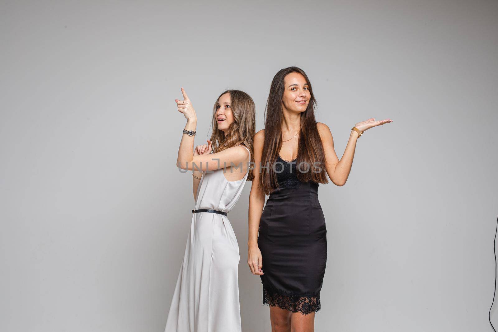 Two pretty women standing in studio pointing their fingers up, isolated on grey background. Party concept. Copy space