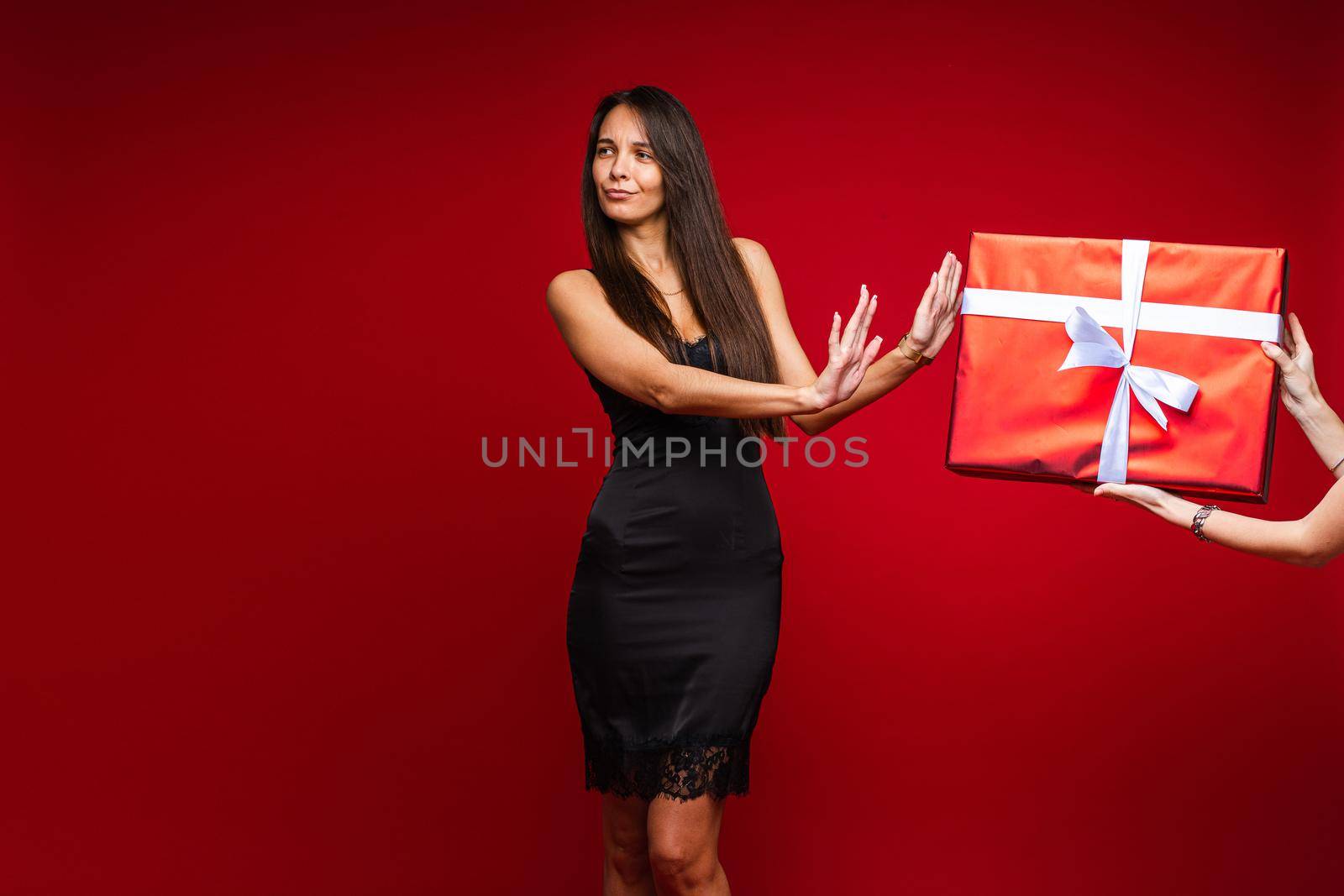 Beautiful young girl in evening black dress refuse from gift on red studio background with copy space for advertising. High quality photo