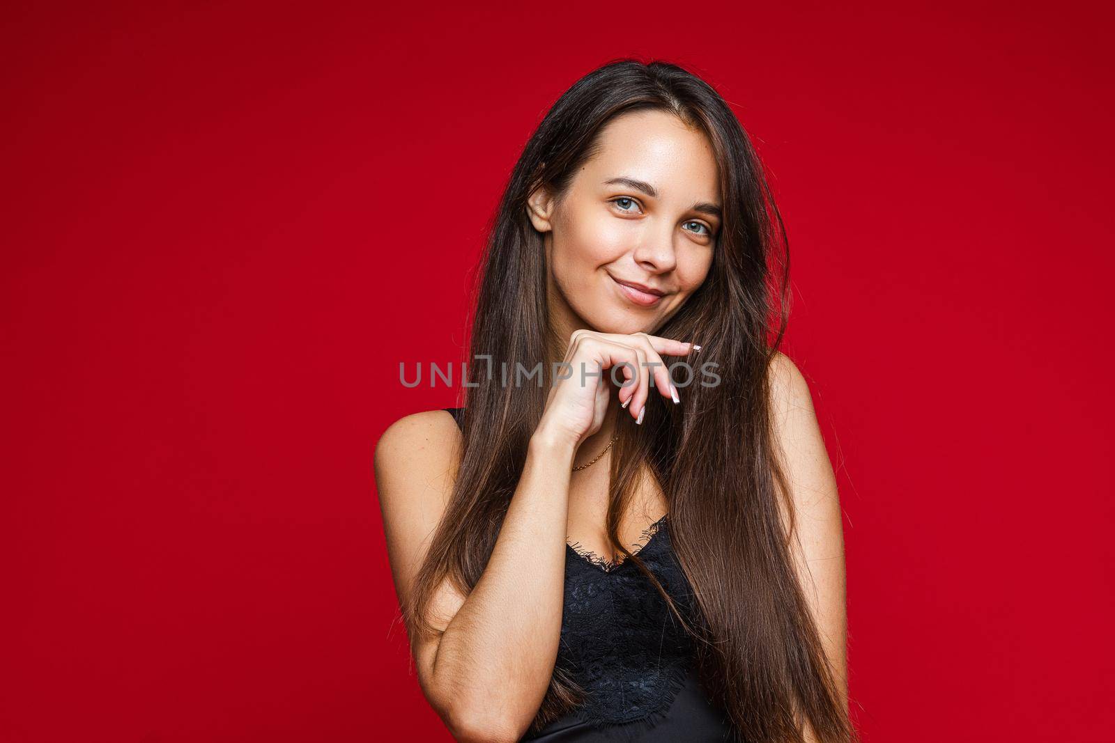 Studio portrait of attractive brunette Caucasian woman in black top with lace smiling at hand at chin. Isolated on red background.