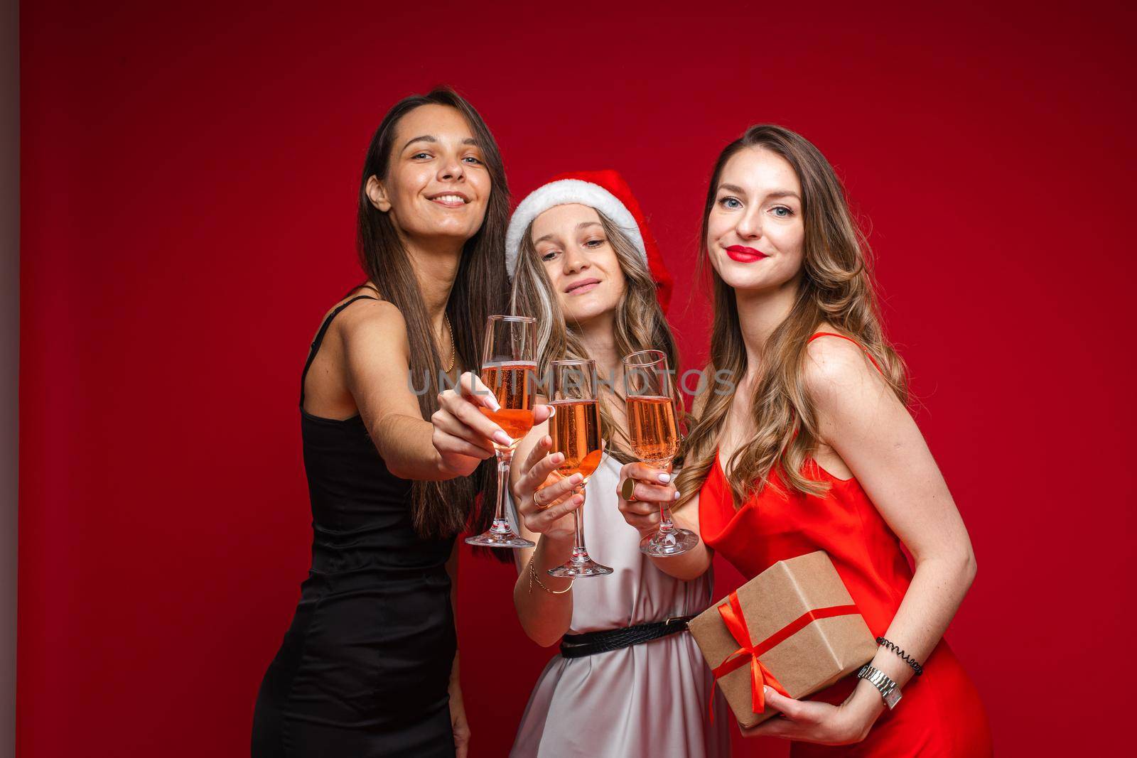 Happy young women friends with gift and champagne celebrating holiday together on party on red background, copy space. High quality photo