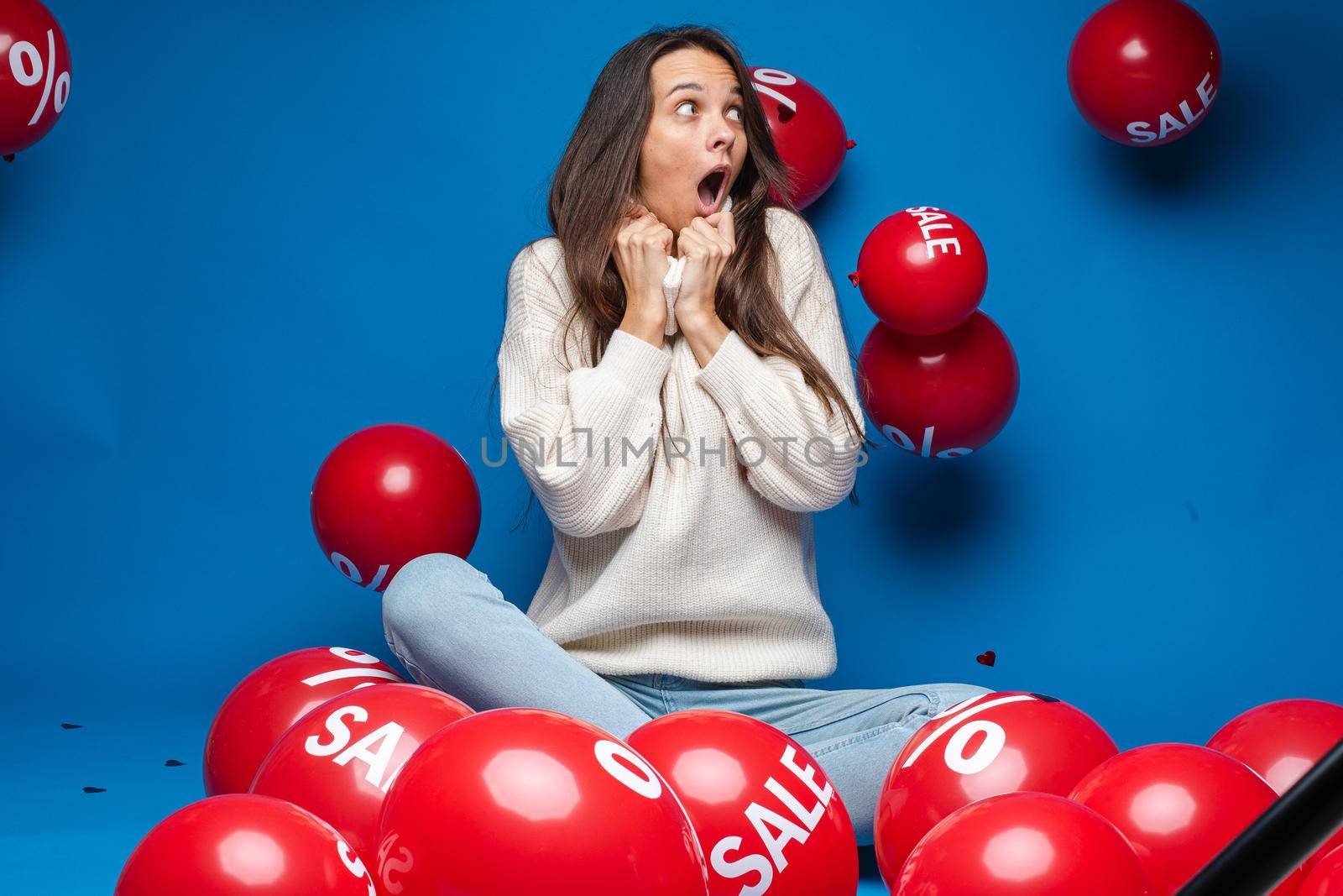 caucasian woman with attractive appearance sits with baloons with percent print and smiles, picture isolated on blue background