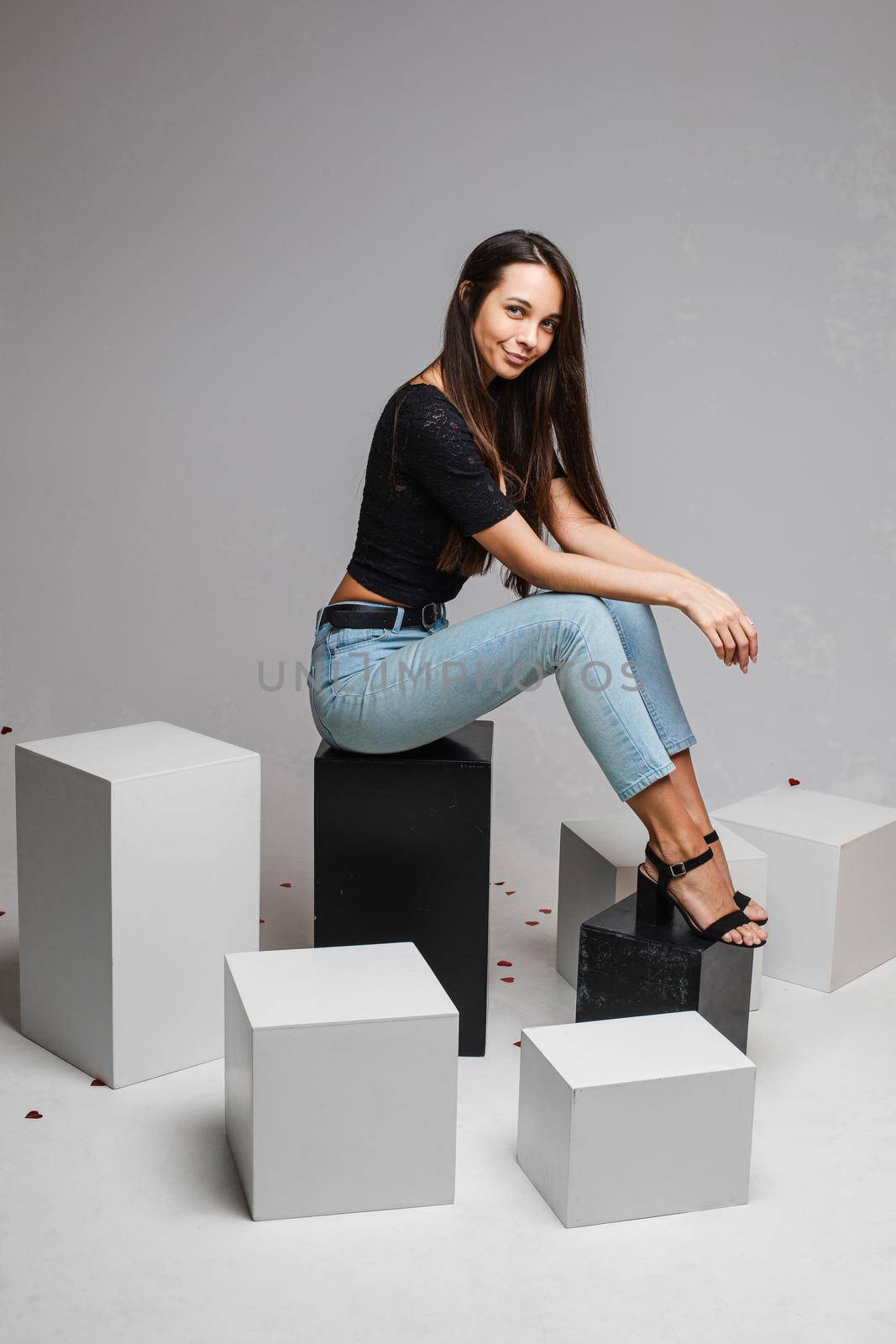 Smiling young fit girl with long hairs posing sitting on black and white cubes on gray studio background with copy space by StudioLucky