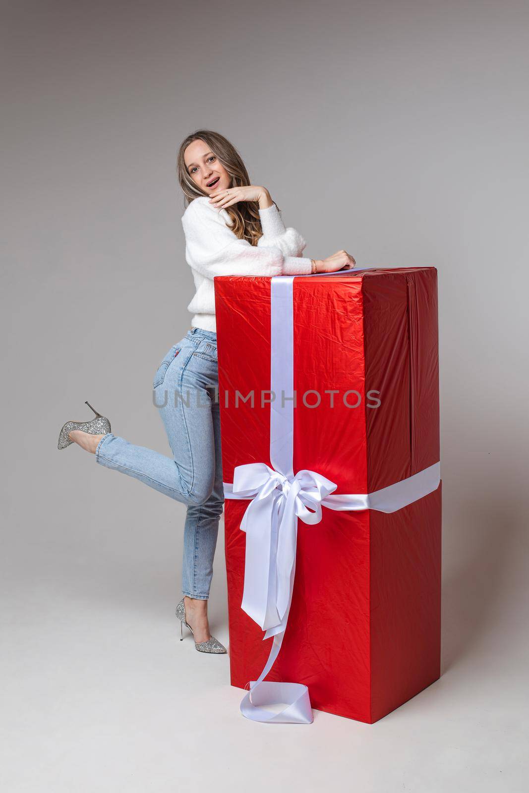 Smiling young woman standing near big red gift box, isolated on grey background. Saint Valentine Day concept