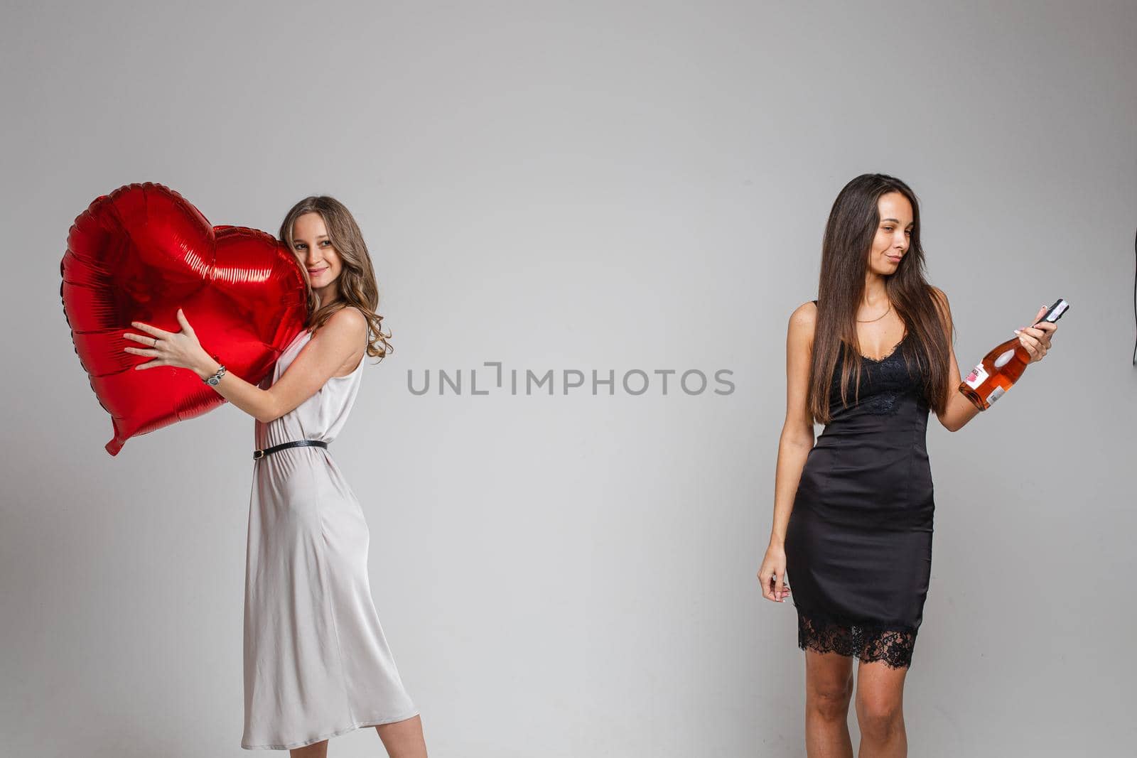 Studio portrait of two friends in silver and black dresses posing on Valentine s day. Fair-haired girl in white dress embracing heart-shaped balloon while brunette in black holding bottle of wine.