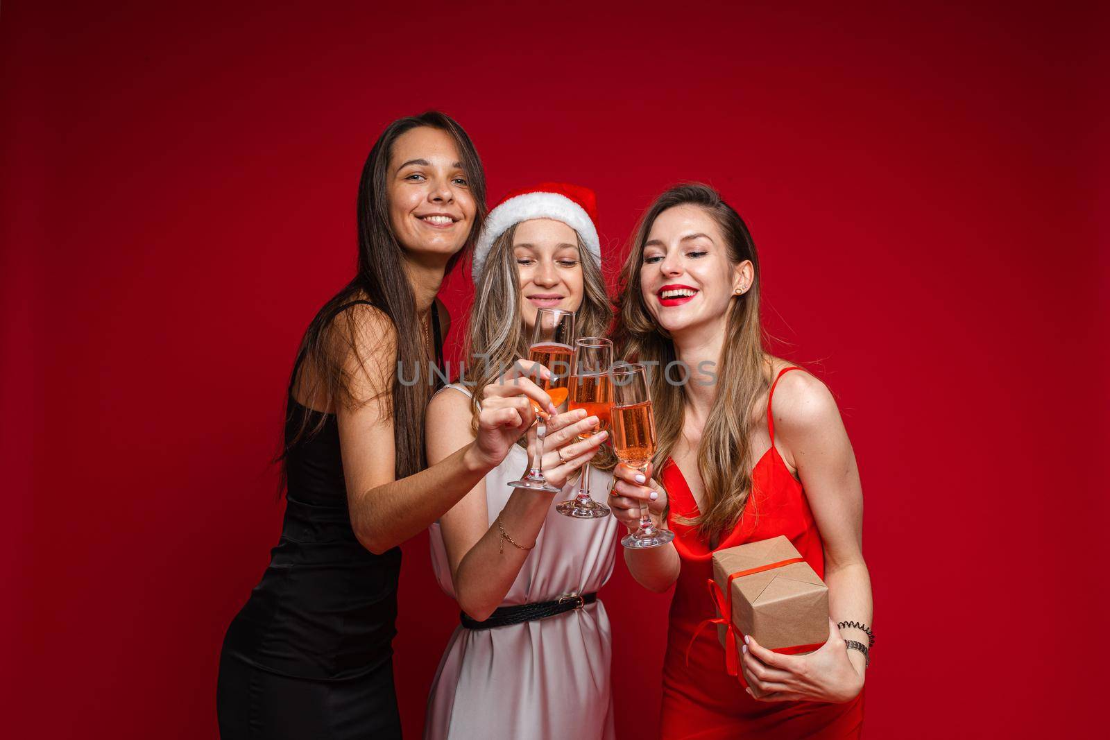 Happy young women friends with gift and champagne celebrating holiday together on party on red background, copy space by StudioLucky