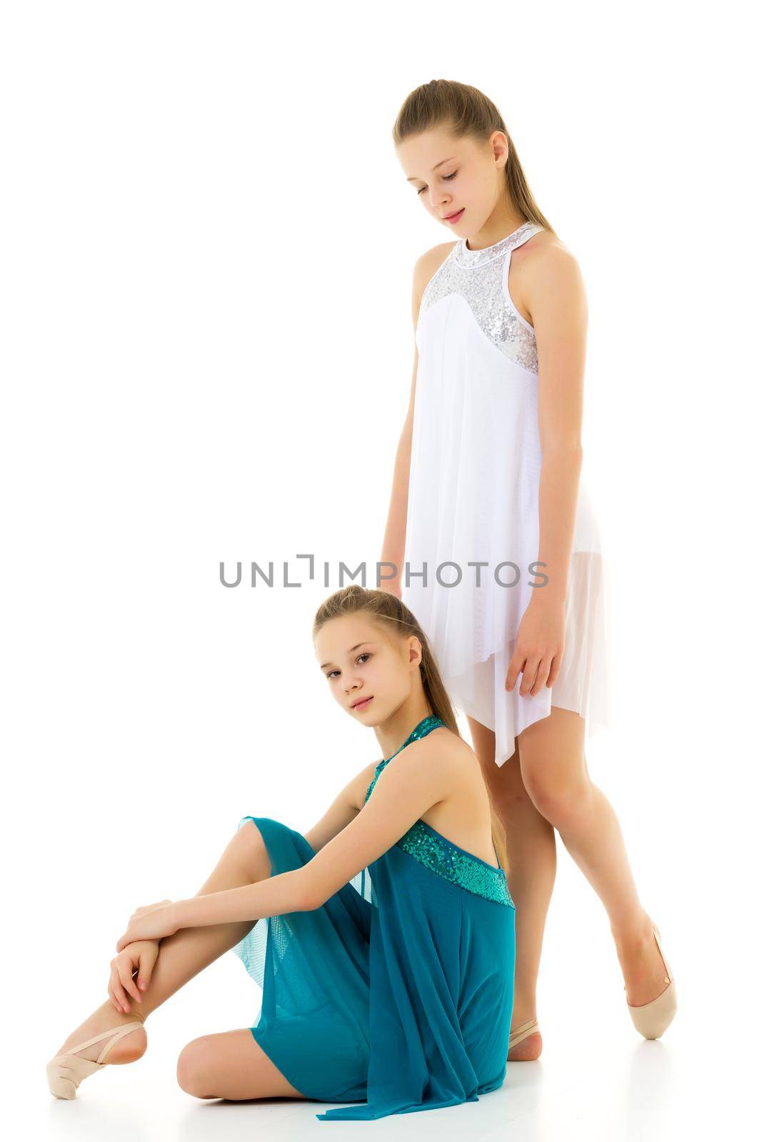 Two Beautiful Teenage Girls in White and Blue Sport Dresses Posing Together Against White Background, Portrait of Pretty Twin Sisters, One of Them Sitting on the Floor, Second Standing Behind