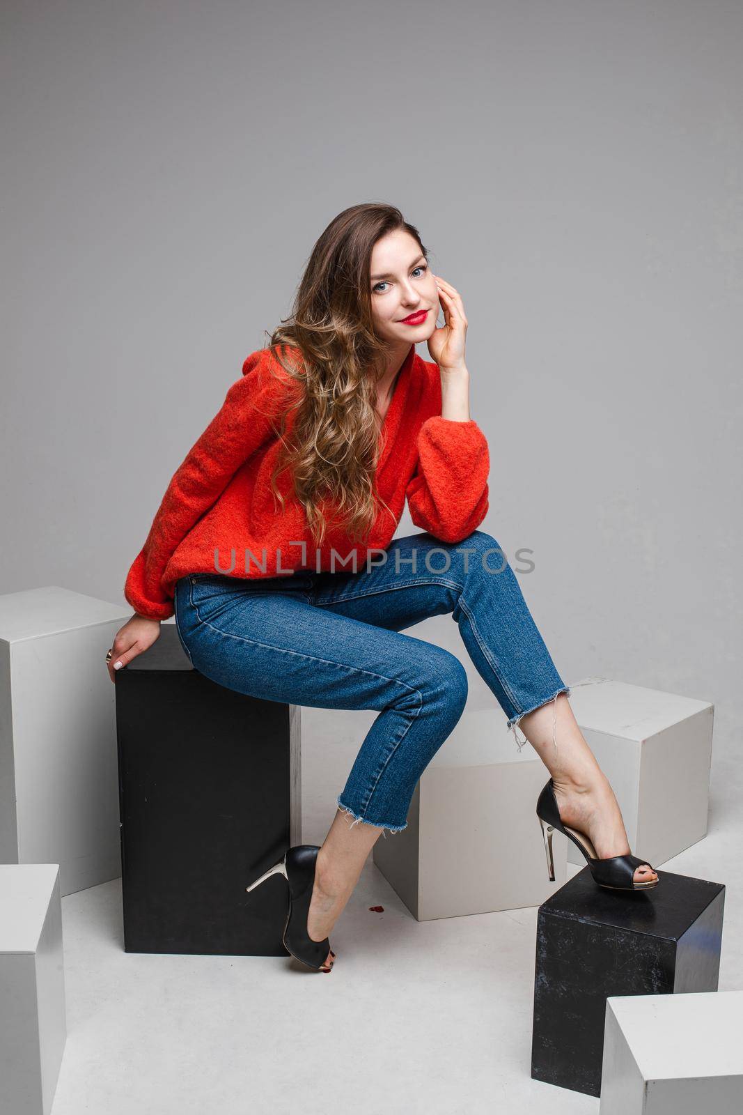 Studio shot of attractive brunette with red lips in stylish casual red pullover and denim jeans and black heels sitting on black and white cubes smiling at camera.