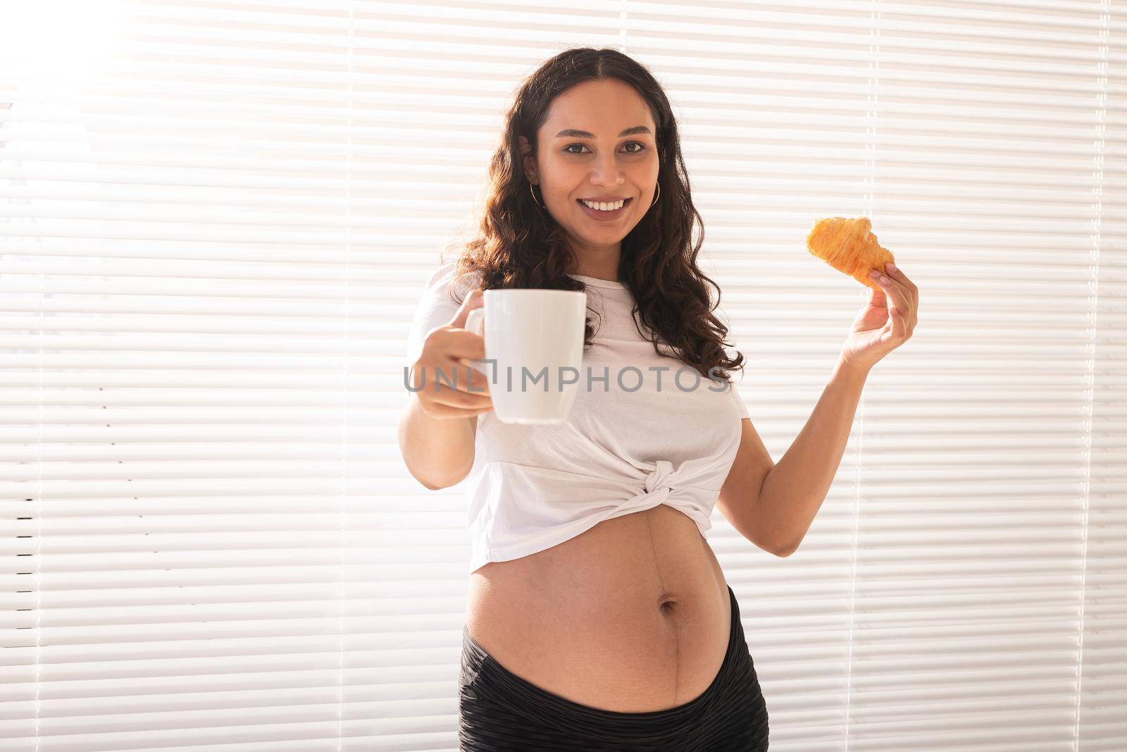 Beautiful pregnant woman holding croissant and cup of coffee in her hands during morning breakfast. Good health and positive attitude while expecting baby