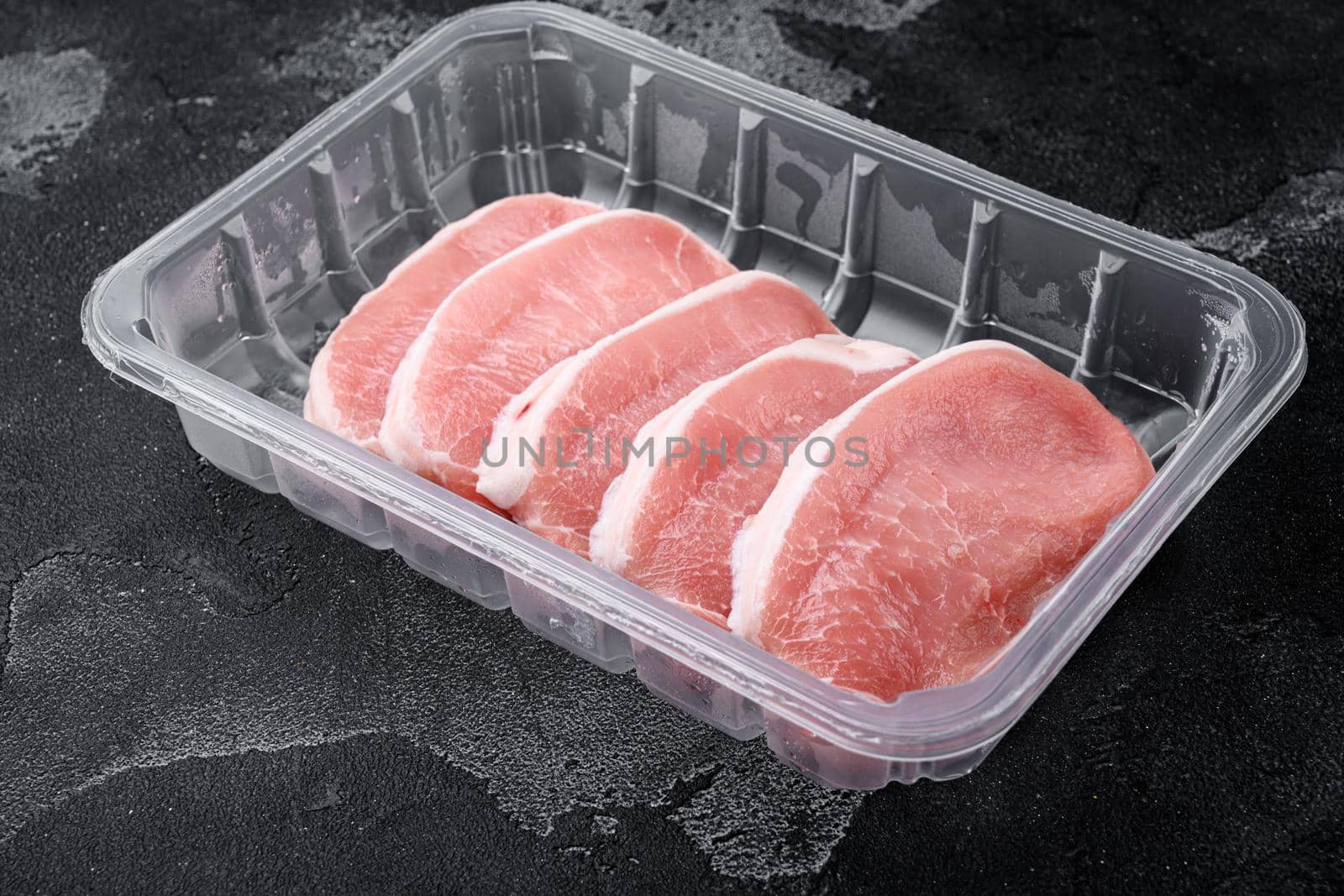 Raw organic meat. Pork steaks, fillets for grilling, baking or frying set, in plastic pack container, on black dark stone table background, with copy space for text