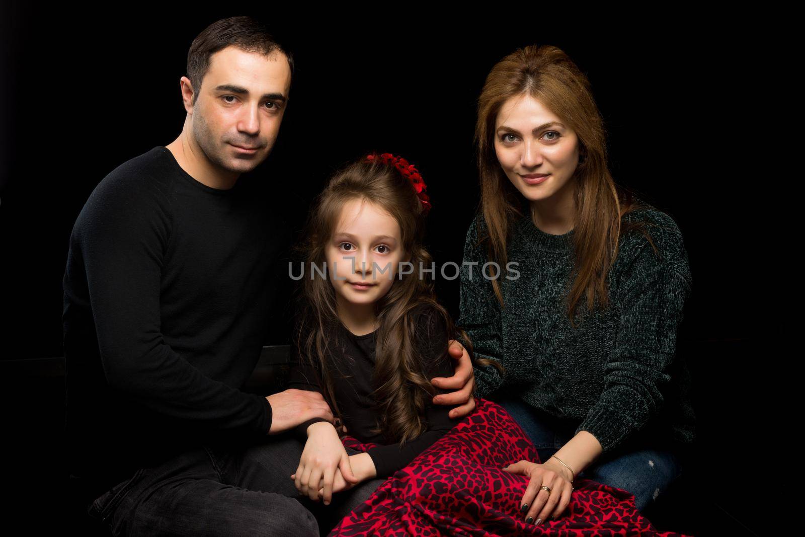 Portrait of Loving Parents Hugging their Adorable Daughter, Mom and Dad Sitting on the Floor and Looking at Their Adorable Girl, Happy Family Sitting Against Black Studio Background