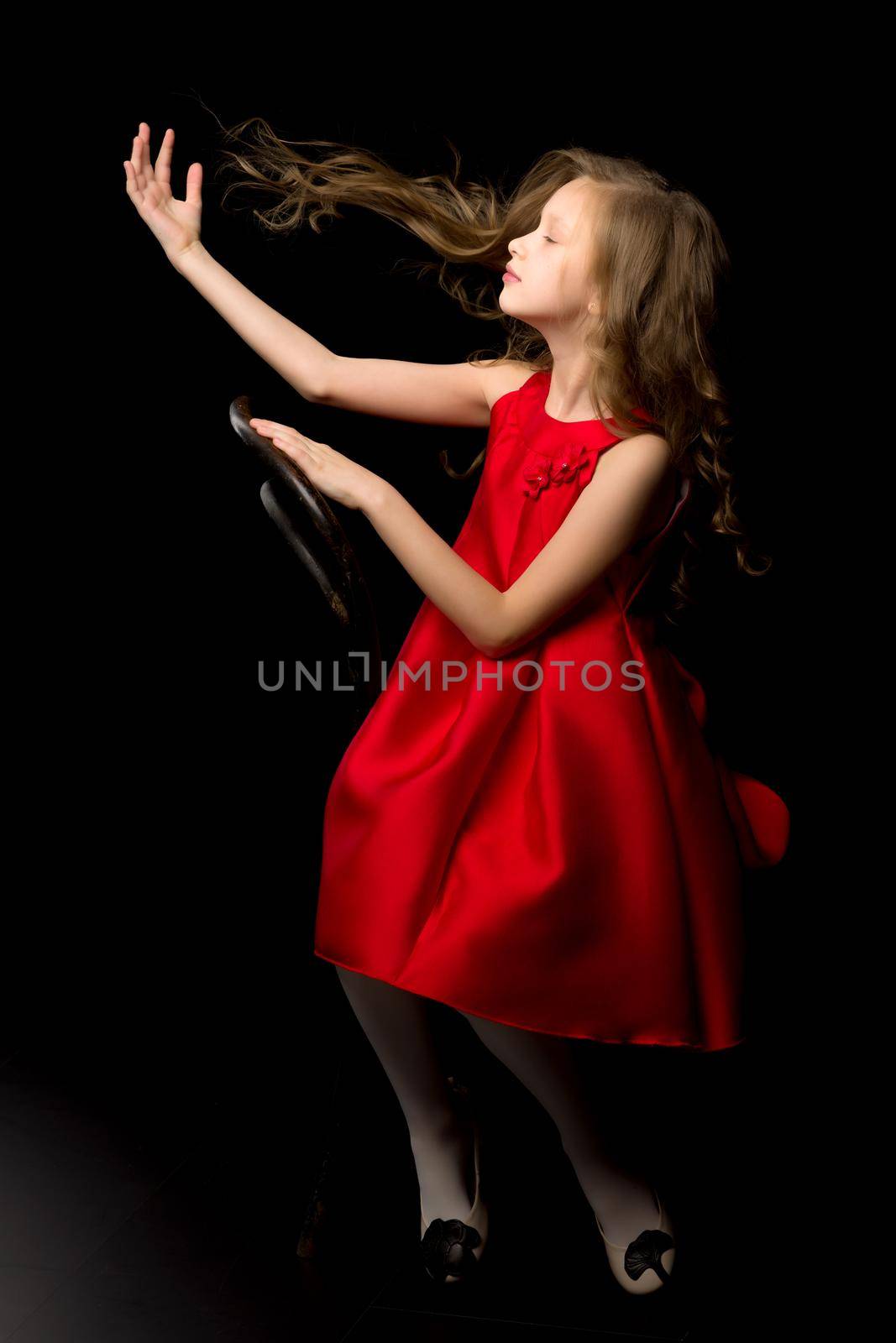 Inspired Beautiful Girl Wearing Red Nice Dress Sitting on Chair Posing at Camera, Portrait of Charming Girl with Flying Long Hair Looking to the Side Against Black Background