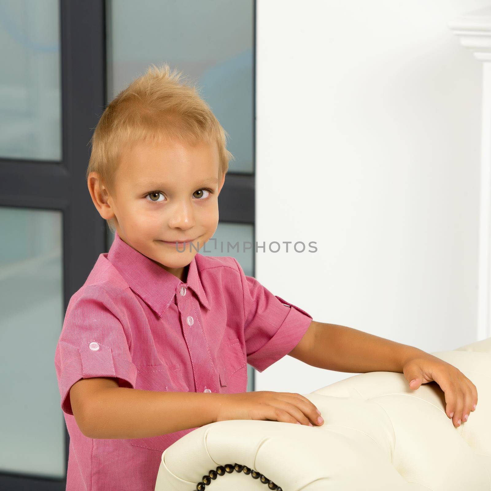Adorable little blond toddler boy near the window and looks indoors. The child looks out the window.