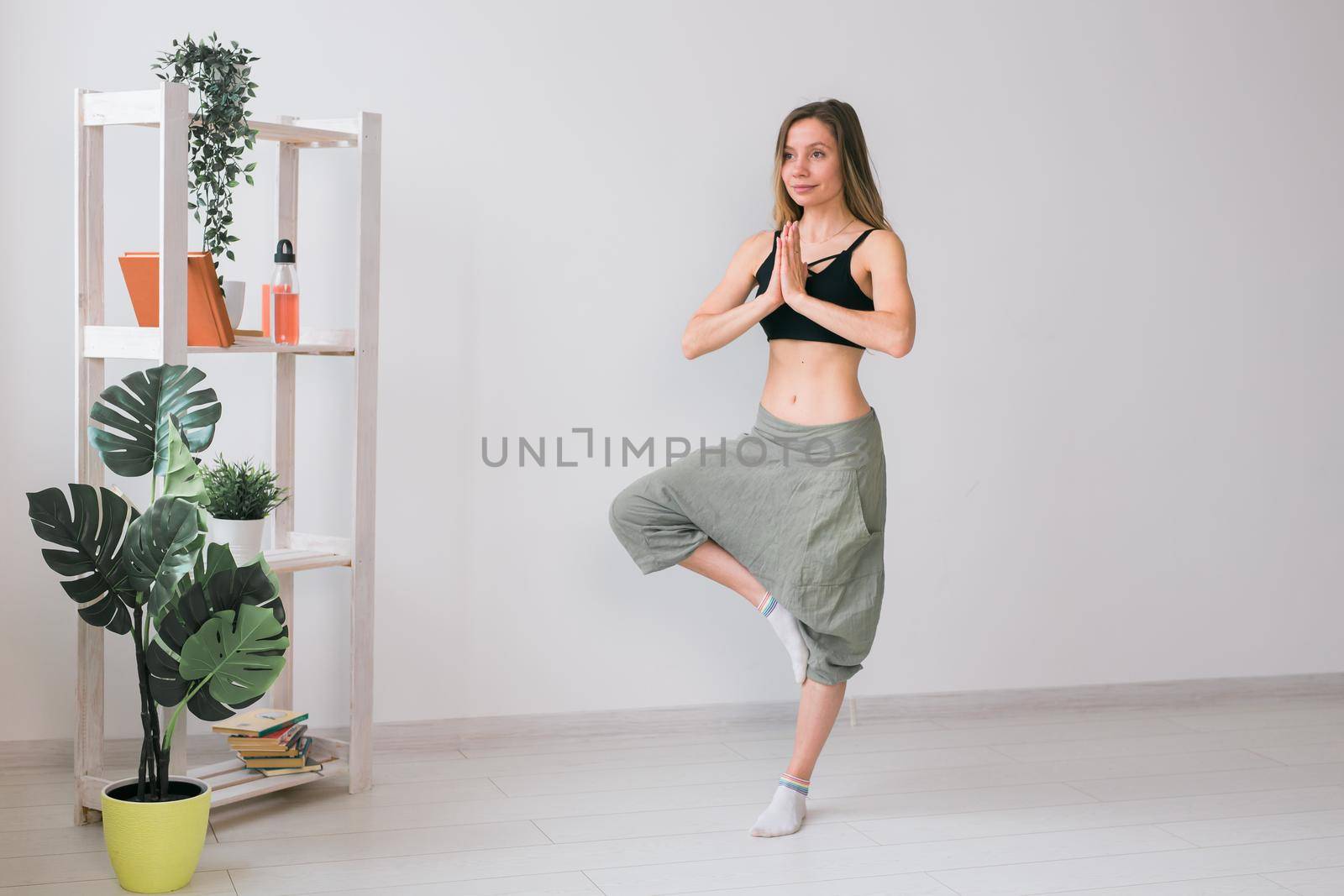 Woman practise yoga performs. Tree pose standing on mat inside of cozy room with plants and greenery. Healthy lifestyle