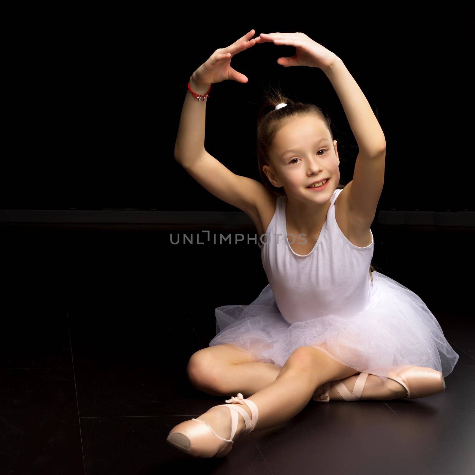 Adorable little girl ballerina. She is posing in pointe shoes, sitting on the floor in the studio. Isolated on a black background.