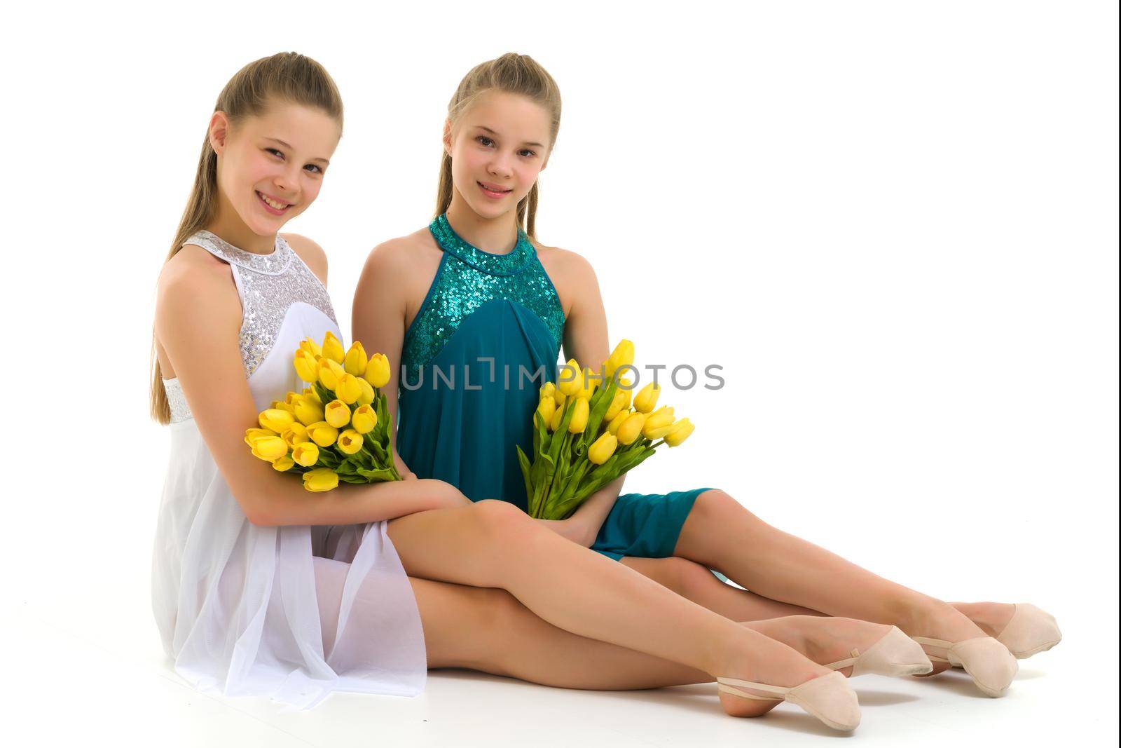 Beautiful girls sitting on the floor with a bouquet of yellow tulips, smiling sisters in summer dresses, portrait of pretty teenage girls on a white background