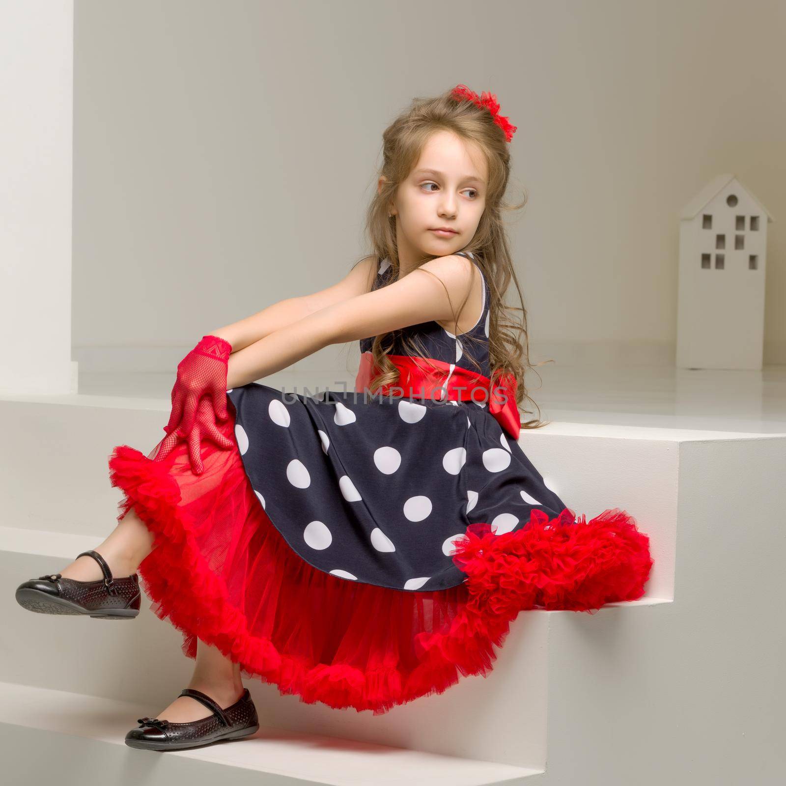 Portrait of Pretty Girl Wearing Blue Polka Dot Dress, Red Gloves and Bow Sitting on White Stairs with Crossed Legs and Looking to the Side, Charming Stylish Girl in Retro Clothes Posing in Studio