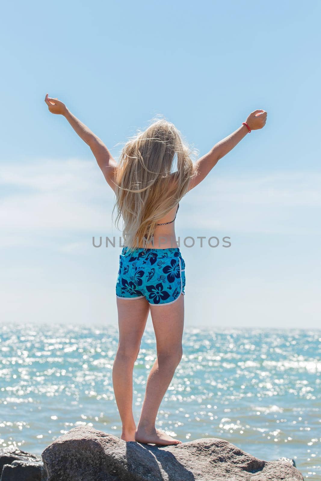 A young blonde girl, a teenager of European appearance in a swimsuit and shorts poses spreading her hands to the sides on seashore against the blue sky and sea water.