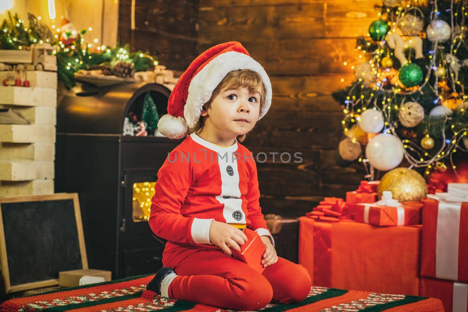 Cute child boy santa with New years gifts on Christmassy background. Elf child. Gifts for winter holidays at fire place