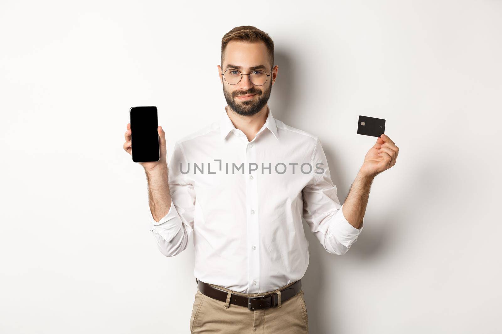 Handsome bearded man showing mobile phone and credit card, shopping online, standing over white background by Benzoix