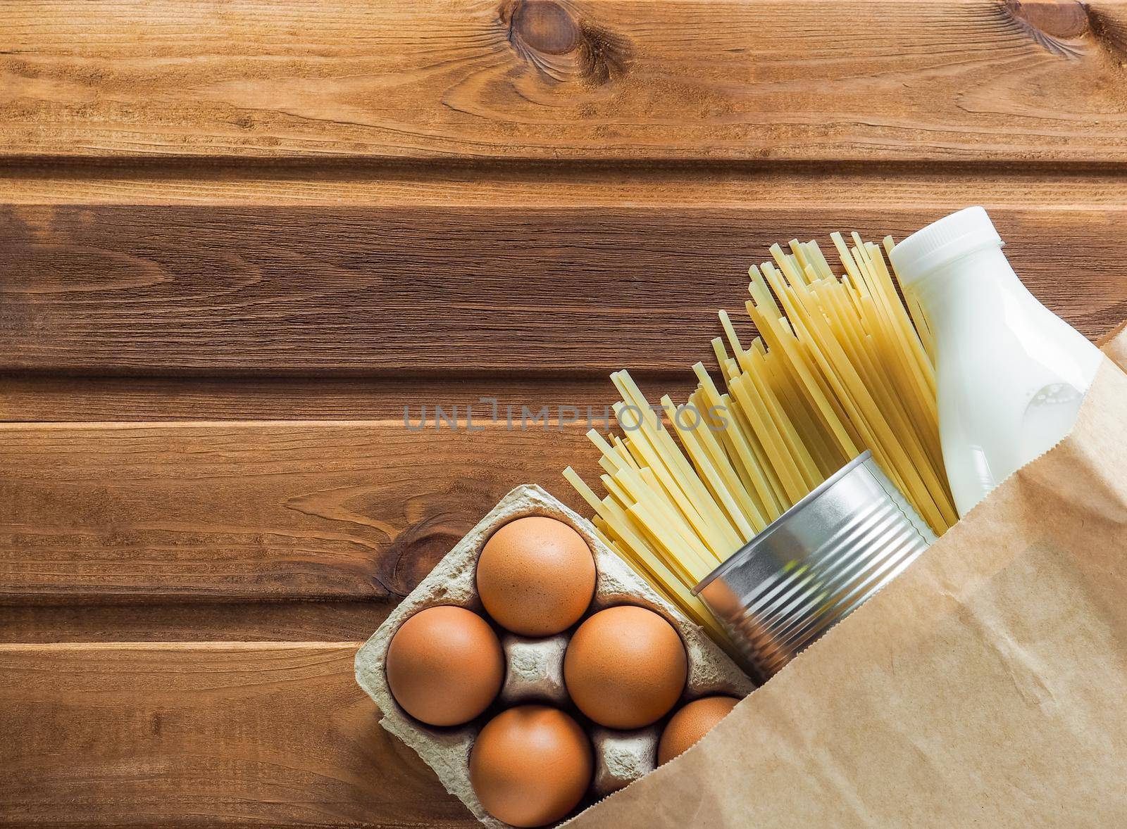 Food delivery, donation, coronavirus, humanitarian assistance. Paper bag with food supplies crisis stock for quarantine isolation period on wooden background. Eggs, milk, canned food, pasta. Copyspace