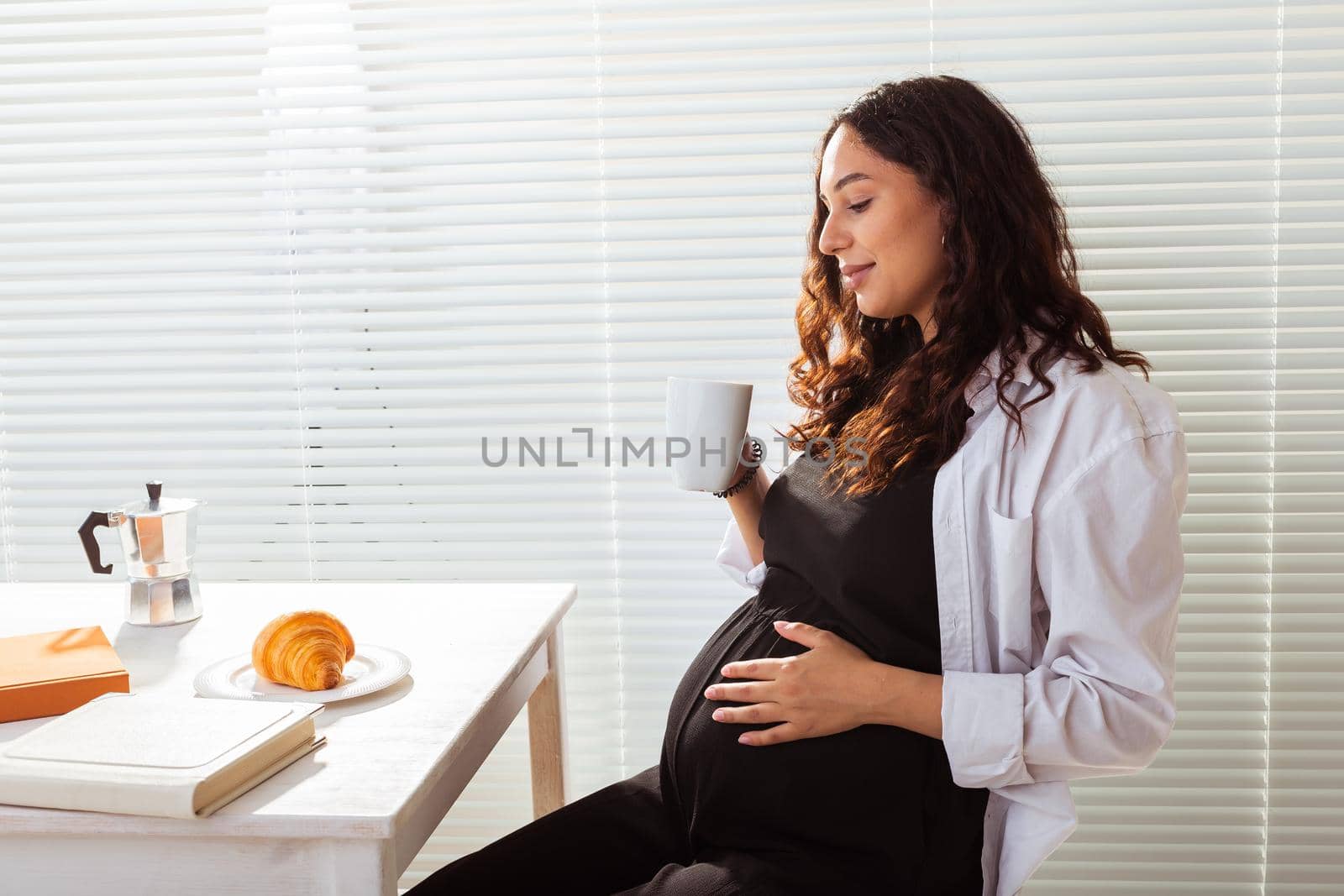 Side view of happy young beautiful woman reading book while having morning breakfast with coffee and croissants on background of blinds. Good morning and pleasant lunch break
