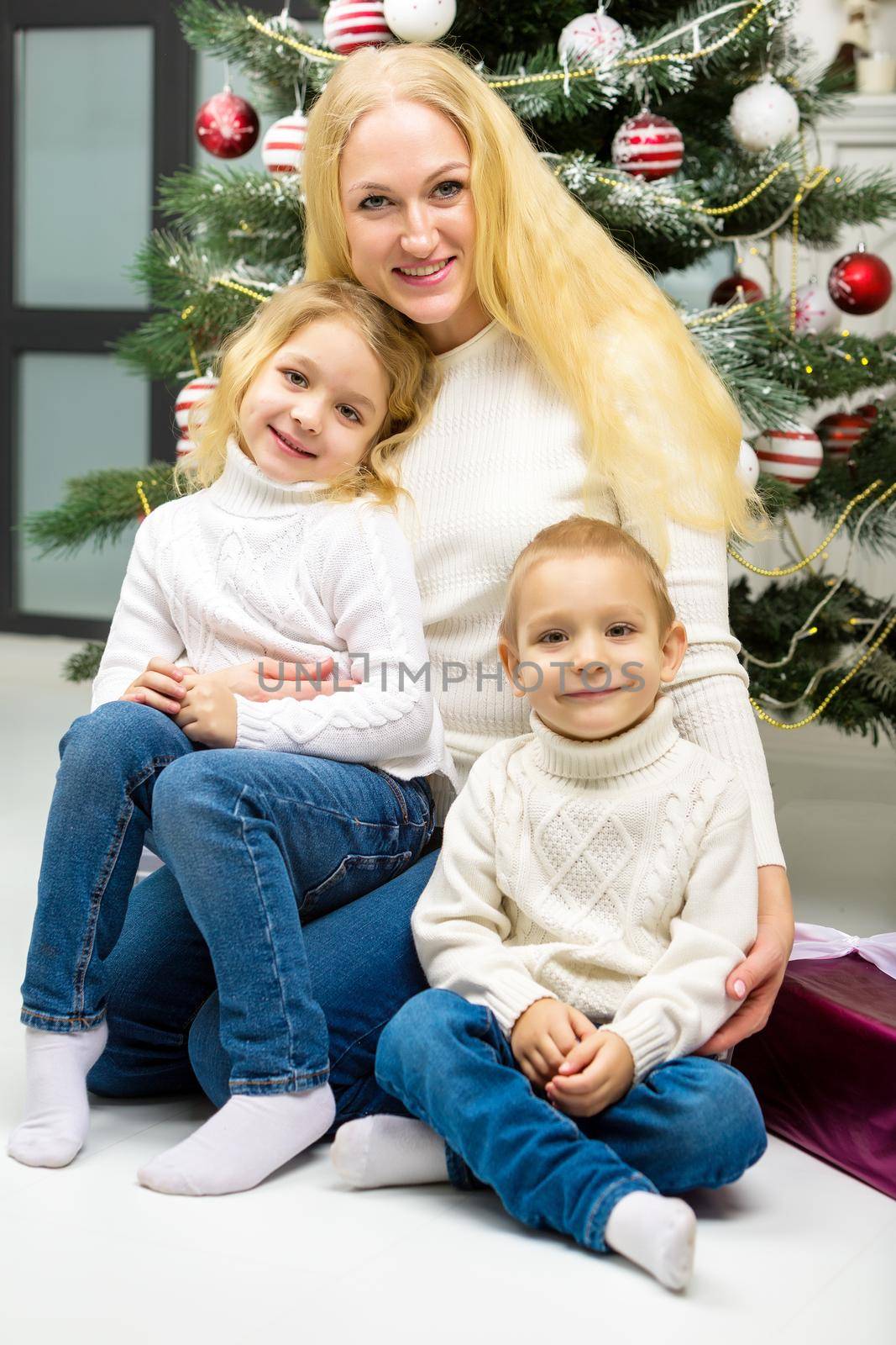 Cheerful Mom Hugging her Cute Little Daughter and Son Happy Family Sitting on the Floor, Smiling Parent and Little Kids Sitting Next Decorated Christmas Tree and Looking at Camera