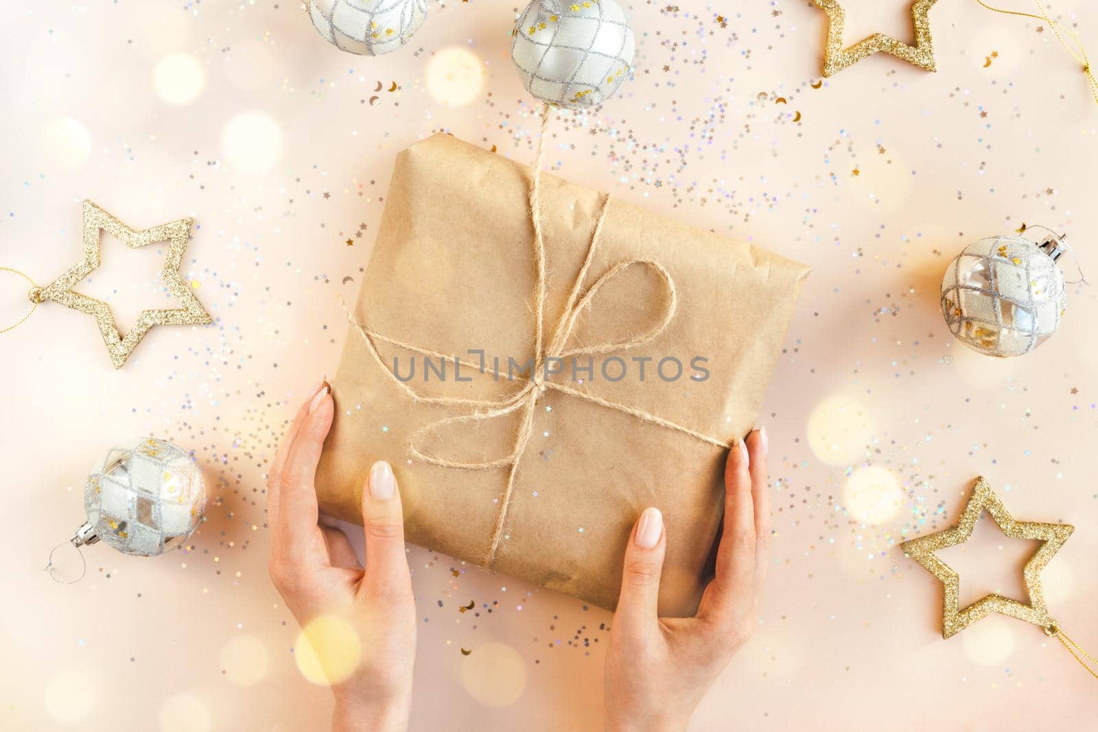 Female hands with natural manicure holding present in kraft paper with jute on soft beige background. Xmas composition. Flat lay. Happy holidays, New Year celebration and giving love concept.