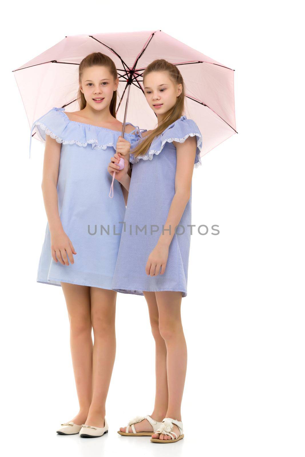 Two Cheerful Pretty Sisters in Identical Light Blue Dresses Standing Under Pink Umbrella, Beautiful Twin Girls in Fashionable Clothes Posing in Studio Against White Background
