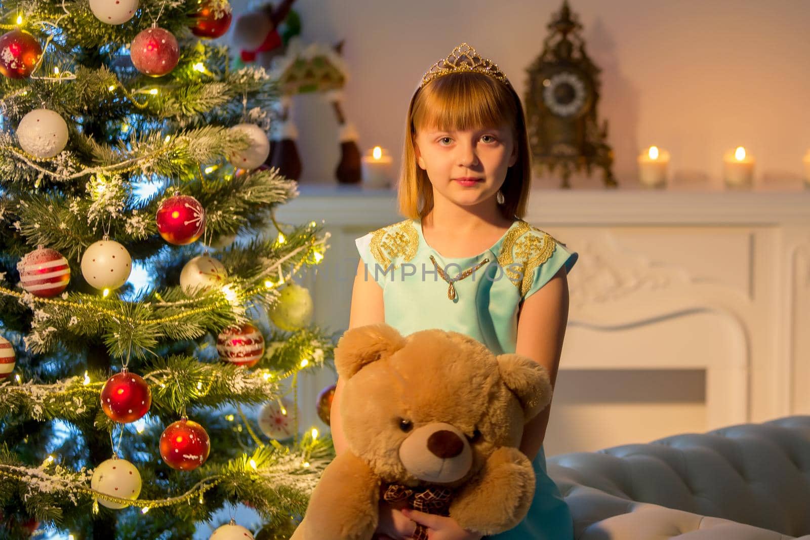 Cute little girl on Christmas night, sitting on a sofa near the New Year trees with a teddy bear in her hands. Family holidays concept.