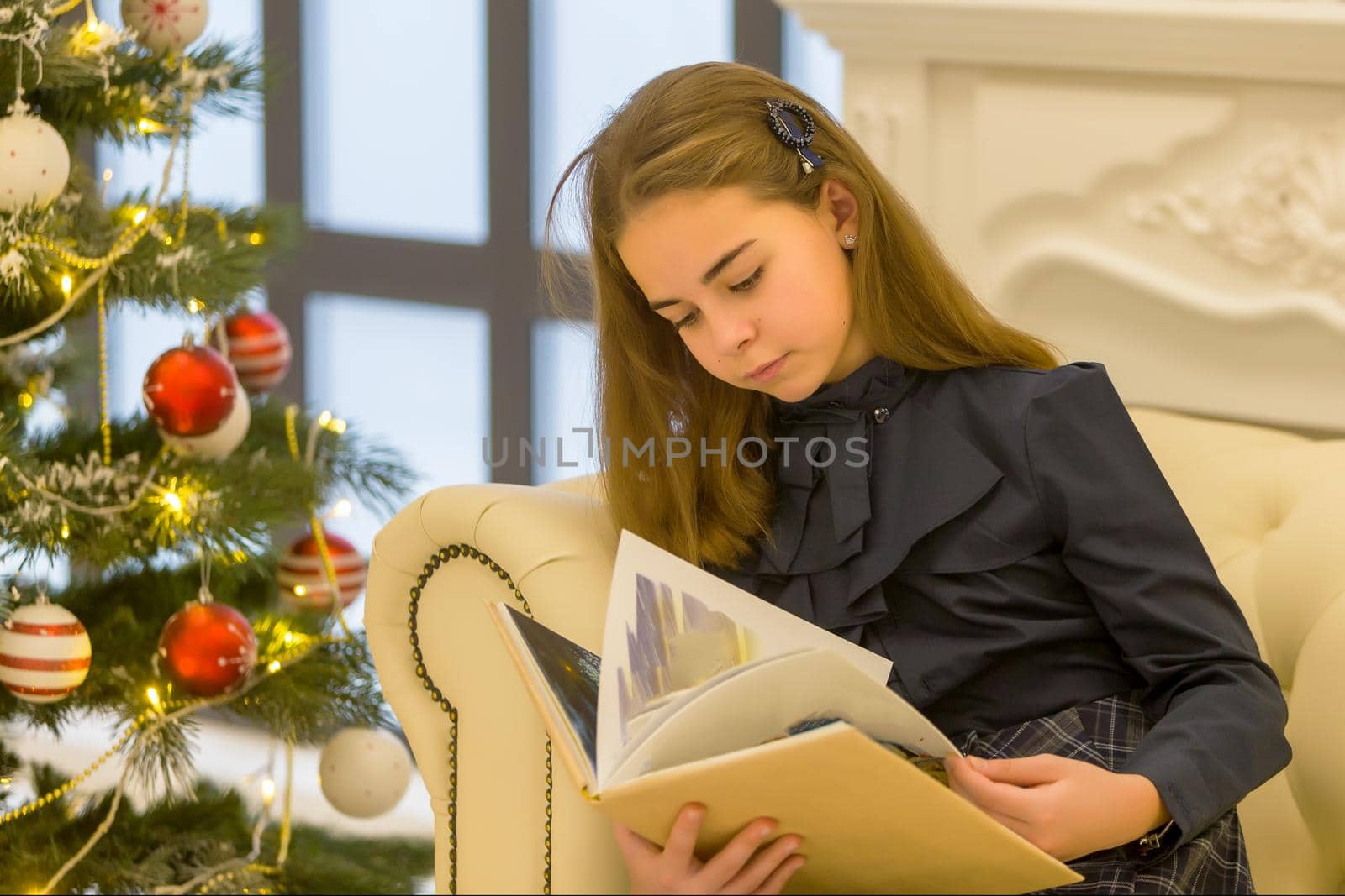Beautiful little girl schoolgirl sitting on a sofa with a book in her hands, near the Christmas tree.