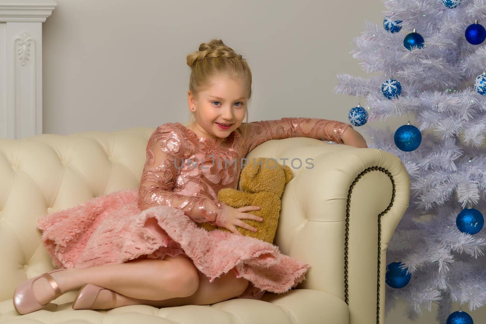 Cute little girl on Christmas night, sitting on a sofa near the New Year trees with a teddy bear in her hands. Family holidays concept.