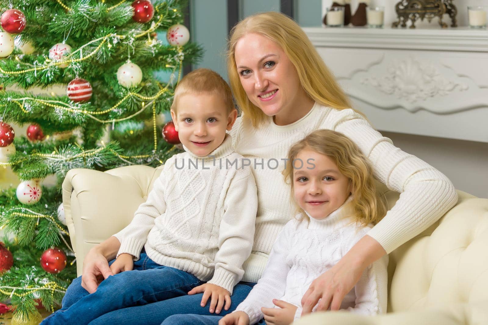 Happy Family Sitting on the Sofa in Christmas Interior with Decorated Fir Tree and Fireplace, Mom Hugging her Cute Little Kids, Her Son and Daughter Looking Lovingly at Her