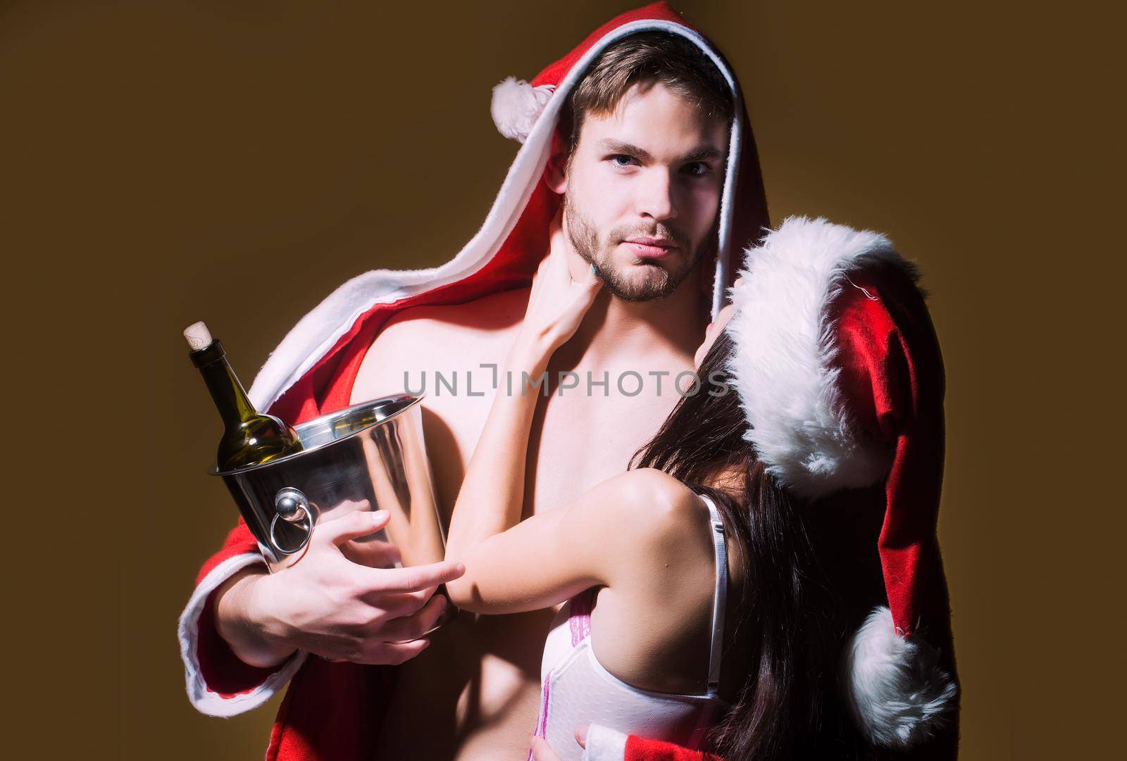 Sexy couple of woman in corset and red christmas hugging muscular man in santa coat.