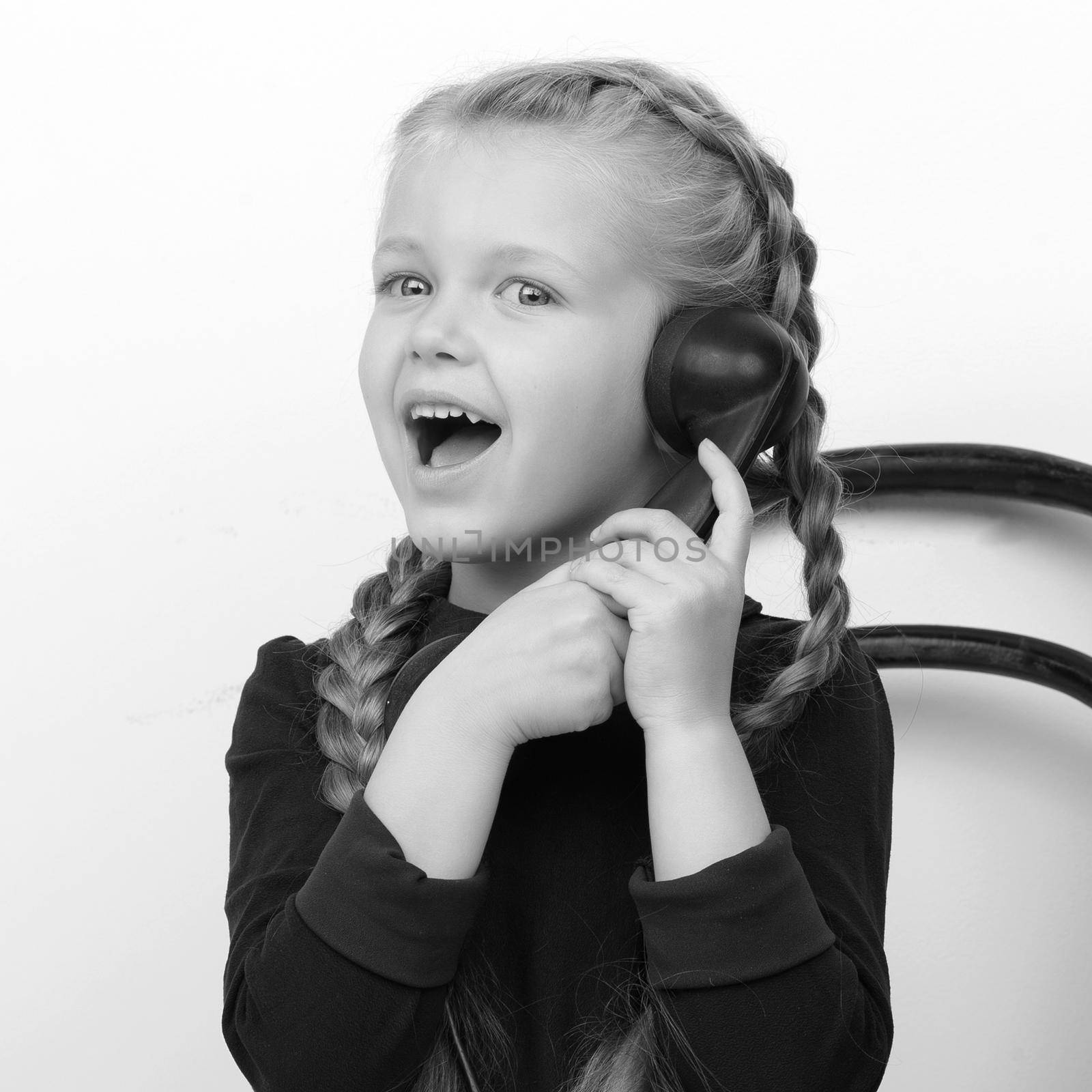 Happy girl talking by old telephone. Black and white portrait of smiling lovely kid sitting on chair. Cute six years old girl with braids speaking on vintage phone