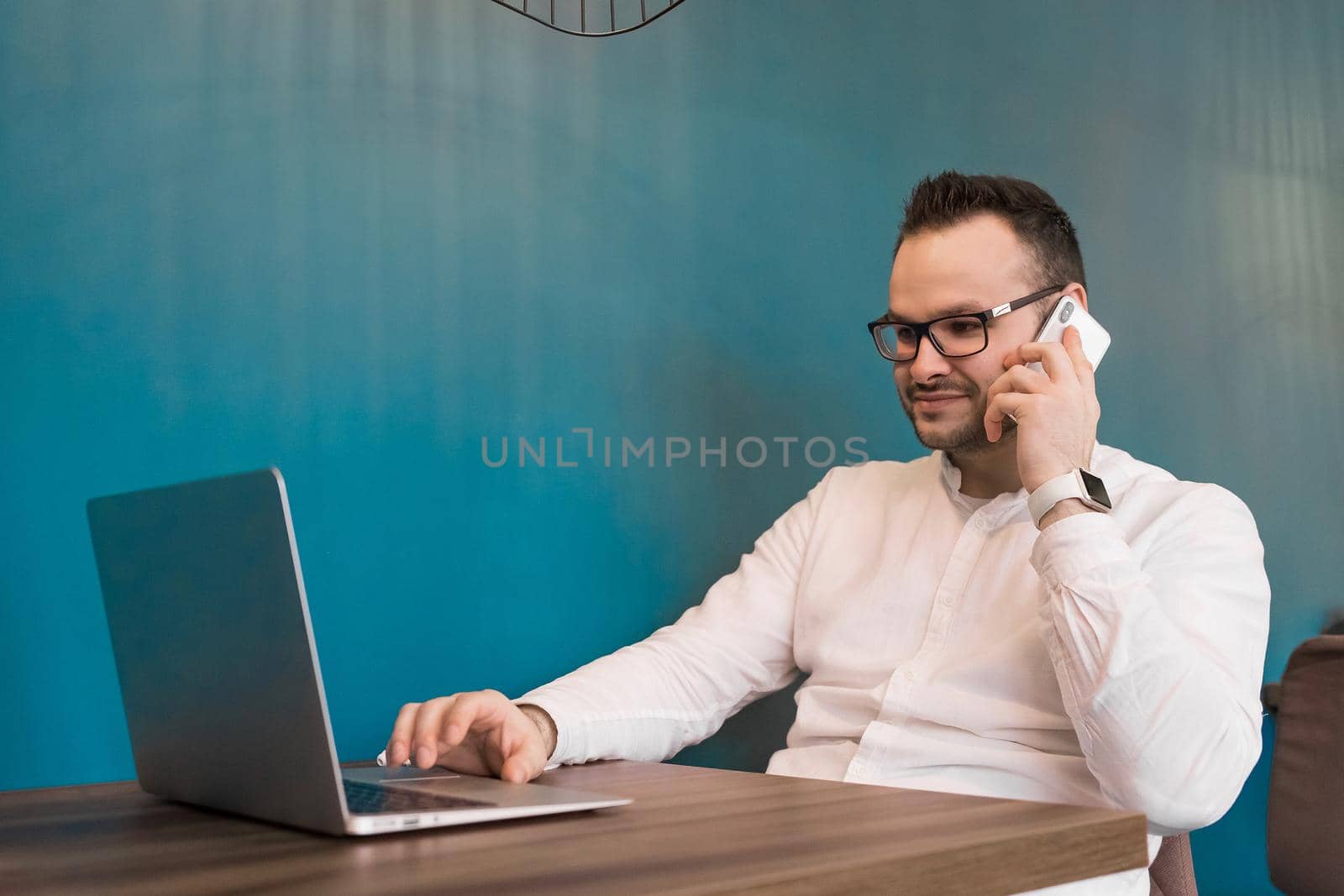 Young businessman in glasses and a white shirt talking on the phone and sitting in a laptop on a blue background.