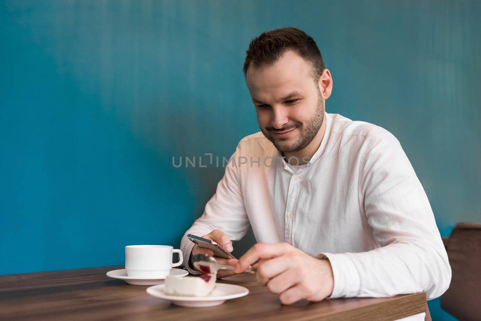 Young attractive businessman in a white shirt sits at a table in the phone and smiles, drinks coffee and eats cake on a blue background.