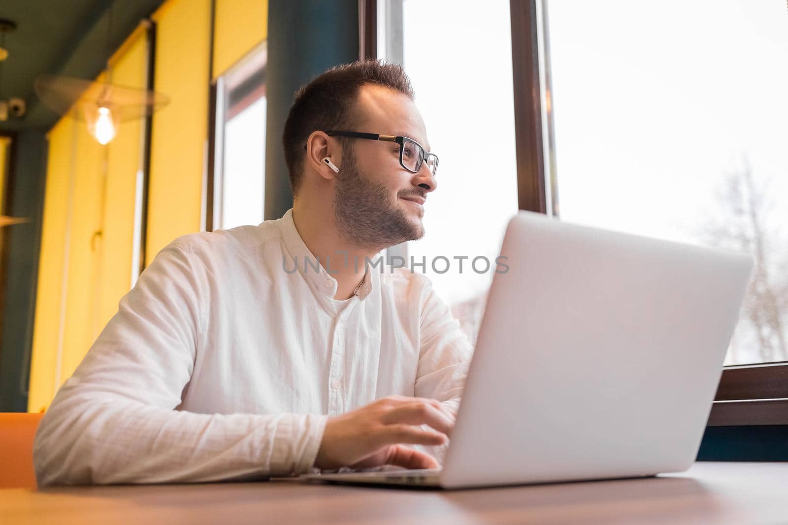 Young handsome businessman in glasses, with a beard, in headphones and a white shirt works in a laptop, looks out the window and smiles.